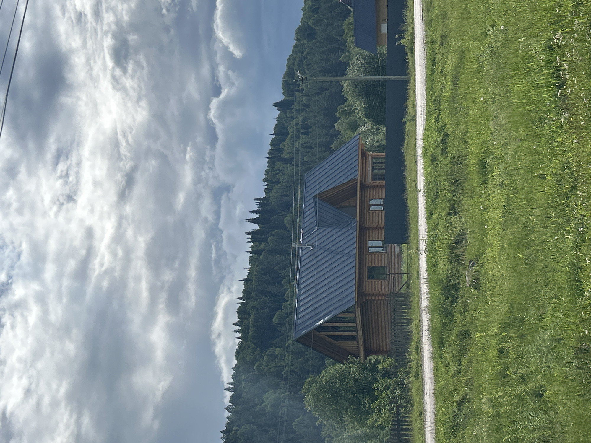 The image depicts a rural landscape with a wooden log house prominently featured in the foreground. The house has a dark, sloped roof and several windows. Behind the house, a dark fence separates it from a lush green forested hill. The sky is overcast with dramatic clouds. In the lower part of the image, a grassy field with scattered wildflowers stretches towards a gravel road. A power line is visible crossing the upper part of the frame. The scene appears to be set in a natural, somewhat remote location. There is no indication of specific people or actions. The location is identified as Usman'gali, Russia.