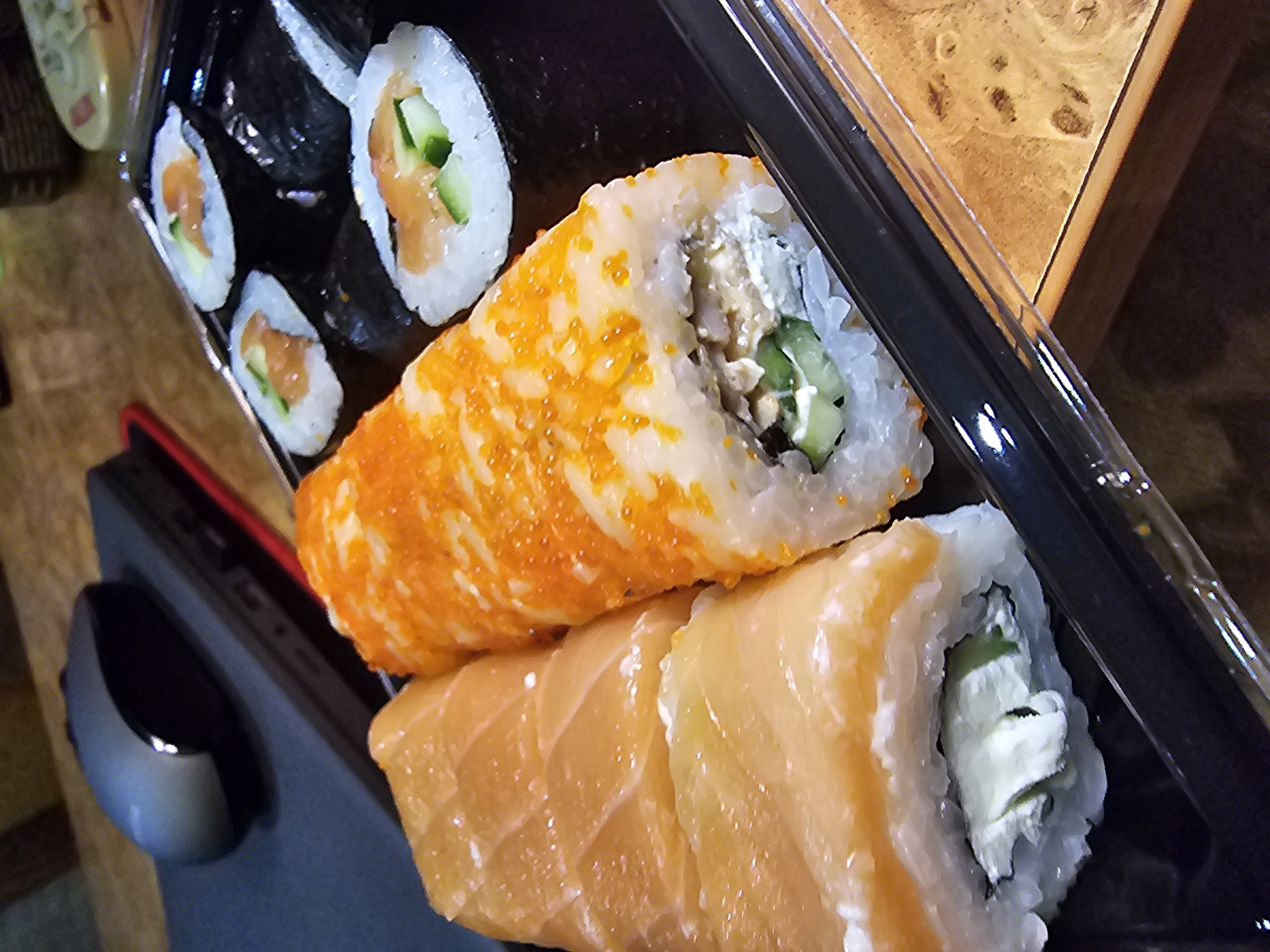 A close-up, high-angle shot shows a black plastic container filled with sushi rolls. On the left side of the container, two large sushi rolls are visible, wrapped in what appears to be raw salmon and filled with cream cheese and cucumber. To the right of these, a roll is covered in bright orange tobiko (flying fish roe) and filled with what looks like a mixture of tuna, cucumber, and possibly cream cheese. Further to the right, three smaller sushi rolls are arranged, each with white rice, nori seaweed, and visible fillings of salmon and cucumber. The container is placed on a wooden surface. In the background, a dark gray computer mouse rests on a dark gray laptop.