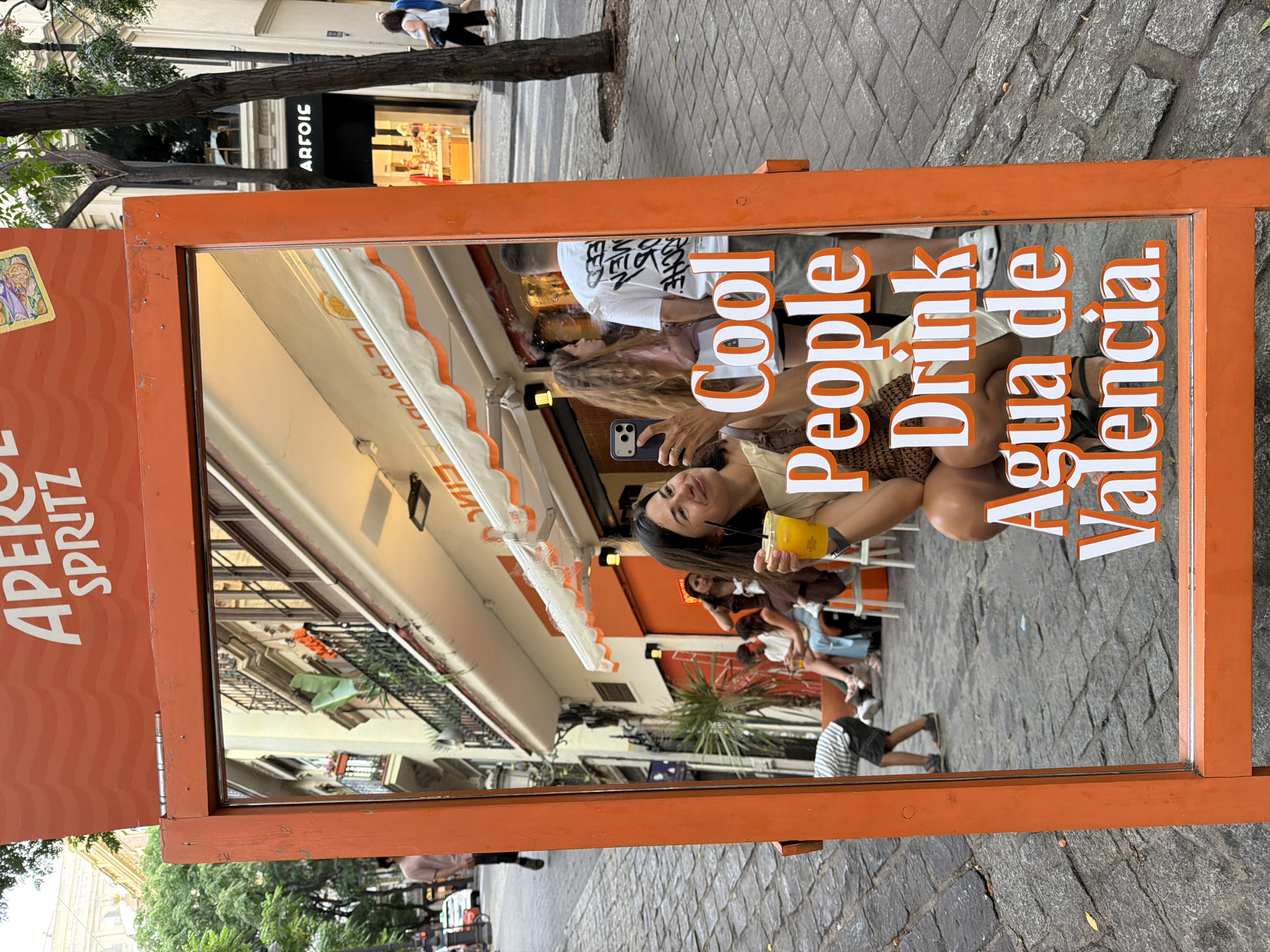 This is a medium-shot, eye-level, outdoor photograph taken in the city of Unknown, Unknown. The primary subject is a woman in the foreground, reflected in a large, orange-framed mirror that serves as a sign. The mirror displays text that reads "Cool People Drink Agua de Valencia." The woman, wearing a patterned top and holding a yellow beverage in her right hand, is captured in a squatting position, taking a selfie with her phone.
In the reflection, other people are visible seated at tables and walking in the background, suggesting a lively outdoor seating area of a cafe or bar. The architecture visible in the reflection includes multi-story buildings with balconies and awnings. The ground surface is paved with cobblestones. The overall atmosphere appears to be a daytime scene in a European urban setting.