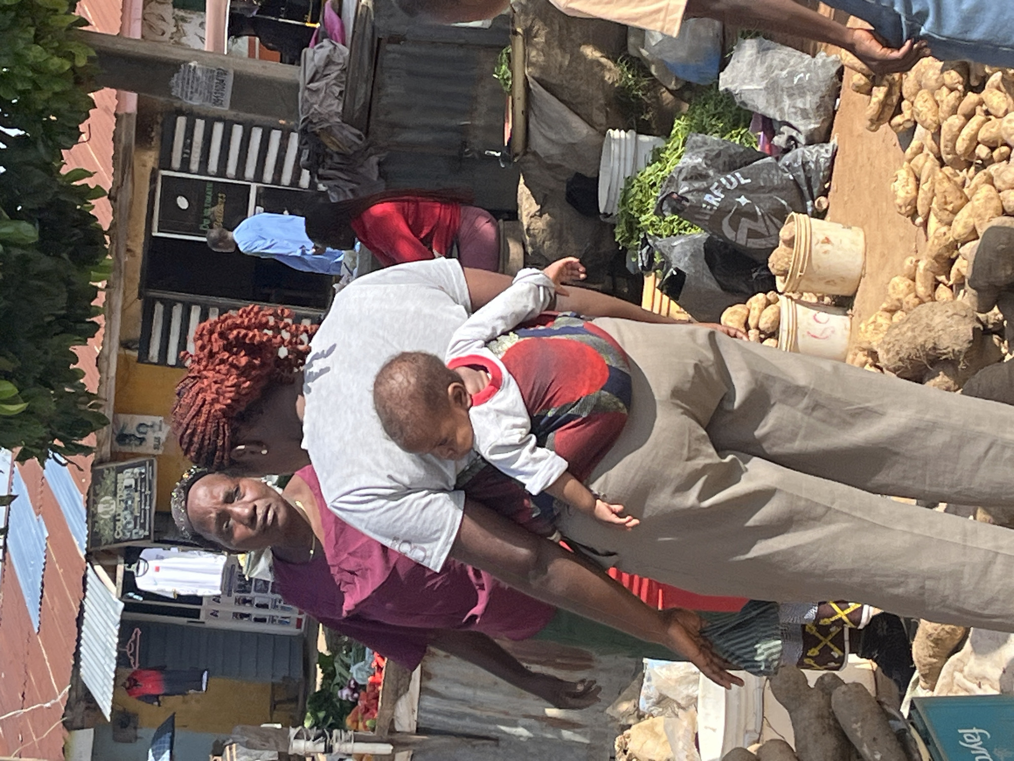 The image depicts a street market scene in Jos, Nigeria. In the foreground, a woman is seen from behind, carrying a baby on her back using a cloth wrap. The baby is wearing a light-colored shirt with red trim. The woman is wearing grey pants. To her left, another woman wearing a dark red top and a headscarf is looking towards the viewer. Behind them, there are market stalls with various goods, including produce displayed on a wooden table and vegetables in sacks and buckets. A person in a light blue garment is visible entering a building in the background. The environment appears to be an outdoor market with corrugated metal roofing on the stalls and a dirt ground. The overall impression is one of a bustling local marketplace.
