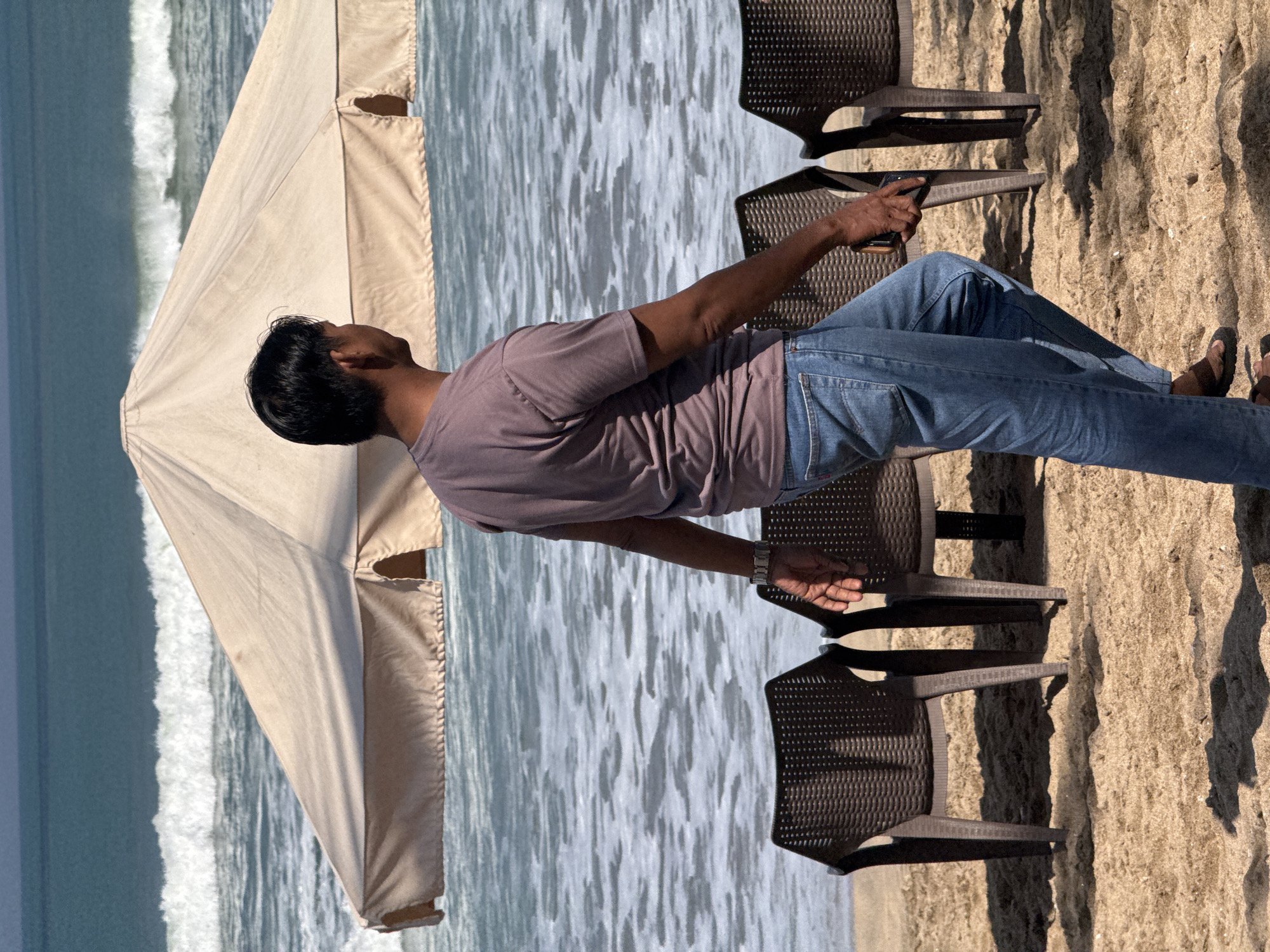 A man wearing a purple t-shirt and blue jeans walks on a sandy beach in Kuta, Indonesia. He is walking away from the camera, and his body is partially obscured by a large, beige umbrella. In the background, the ocean is visible with gentle waves. In front of the umbrella and behind the man, there are three dark plastic chairs arranged on the sand. The man is wearing a watch on his left wrist and appears to be holding a phone in his right hand. The lighting suggests it is daytime.