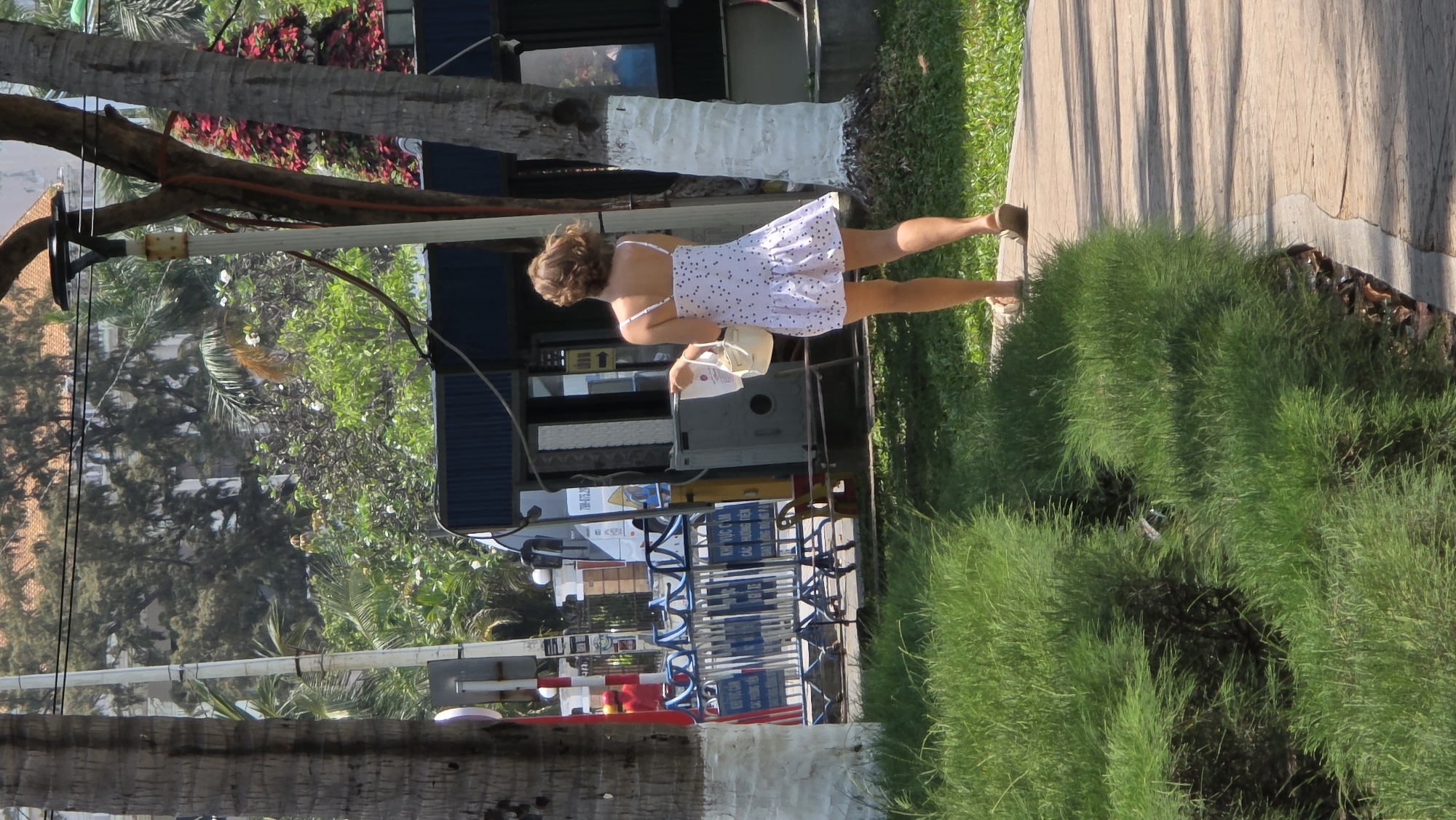 A woman walks on a paved path next to green vegetation in Nha Trang, Vietnam. She wears a white dress with dark polka dots, brown sandals, and carries a white purse. A tree trunk stands beside the path. In the background, there is a refueling station with visible signs in Vietnamese. Other trees and greenery are also present.
