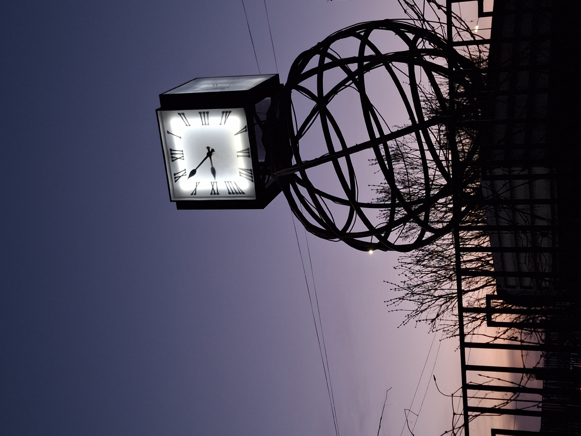 A low-angle, rotated image captures a square clock illuminated against a dusky sky, indicating it's late afternoon or early evening in Chaikovsky, Russia. The clock face displays Roman numerals and the hands suggest the time is around 5:35. Attached to the clock is a decorative metal sphere composed of interlocking rings. To the right, silhouettes of bare tree branches and a fence add depth and context to the urban landscape. The sky transitions from a deep blue in the upper left to a softer, purplish hue with hints of orange near the horizon on the right side, further suggesting the setting sun. The image is tilted approximately 90 degrees.
