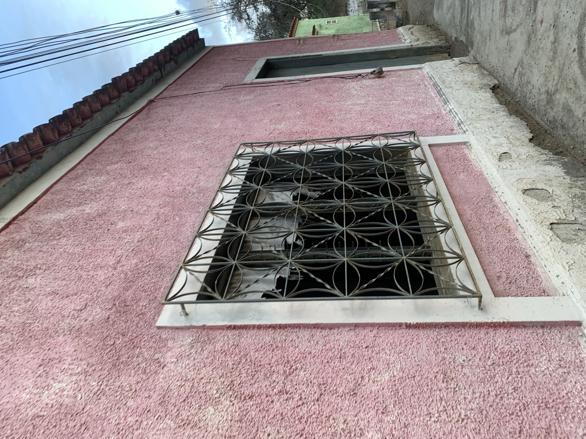 The image shows a side view of a pink stucco building with a distressed white base. A decorative metal grate covers a window. To the right of the window, a dark green door is visible. The building has a tiled roof. The ground in front of the building is a concrete pathway with some dirt and debris. In the background, another building painted green can be seen, along with trees and utility wires. The sky is cloudy. The scene appears to be located in a residential area of Santa Ana, Venezuela.
