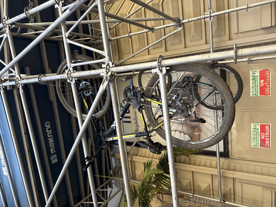 Two bicycles are suspended from scaffolding. One bicycle is black and yellow, while the other has a teal accent color. The scaffolding obscures portions of both bikes. The background features a light-colored building with repeated architectural patterns and some greenery visible below. Warning signs reading "Salida de Emergencia - NO OBSTRUIR" are attached to the building facade. The image is oriented at an angle, making the scene appear tilted.