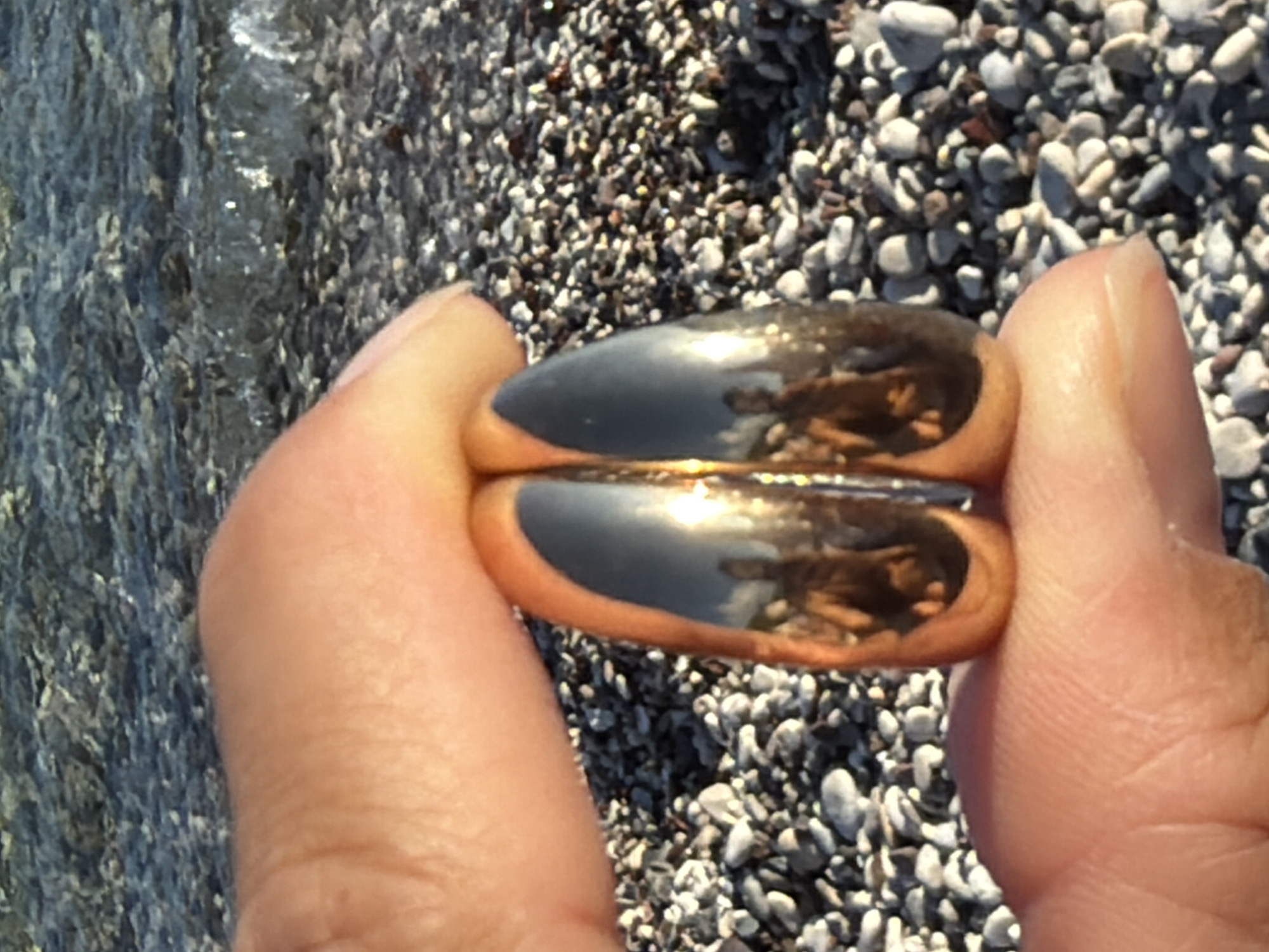 The image depicts a close-up view of a hand holding two gold rings against a backdrop of a pebble beach and the sea. The water is clear, with gentle waves lapping onto the shore, suggesting a coastal location. The pebbles on the beach are varied in size and color, predominantly gray and white. The rings are highly polished, reflecting the sunlight and the surrounding environment, including what appears to be a reflection of a person's face or upper body on their surface. The hand holding the rings is positioned in the foreground, with the thumb and index finger forming a cradle for the jewelry. The lighting suggests it is daytime, possibly late afternoon due to the warm glow of the sun. The overall impression is a serene, natural setting, likely a beach. The image does not contain any elements that would specifically indicate the city of Budva, Montenegro.