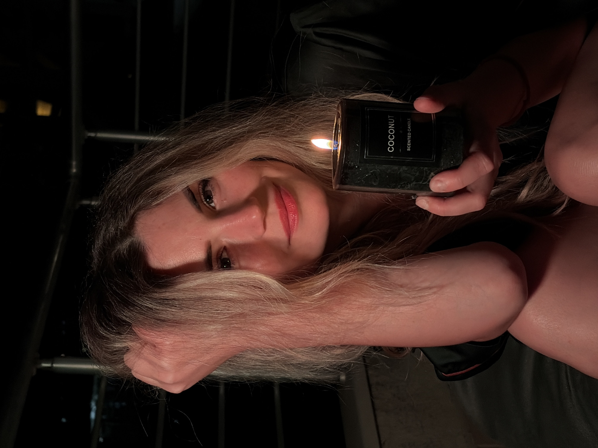 A young woman with blonde hair, wearing dark silk attire, is captured in a medium close-up, holding a lit coconut-scented candle in her right hand. Her left arm is raised, with her hand resting on her head, partially obscuring her face from the left side. The woman is looking at the camera with a slight smile and her lips are painted with a vibrant red lipstick. The candle, a dark cylindrical object with a white label that reads "COCONUT SCENTED CANDLE," is held at chest level. The background is dark, suggesting an outdoor setting at night, possibly on a balcony or terrace, with faint lights visible in the distance, implying an urban environment. The railing of a balcony is visible in the upper portion of the image. The lighting is dim, with the primary light source being the candle flame, casting a warm glow on the woman's face and hand. The scene suggests a relaxed and intimate moment. The location of Budva, Montenegro, cannot be determined from the visual information present in the image.