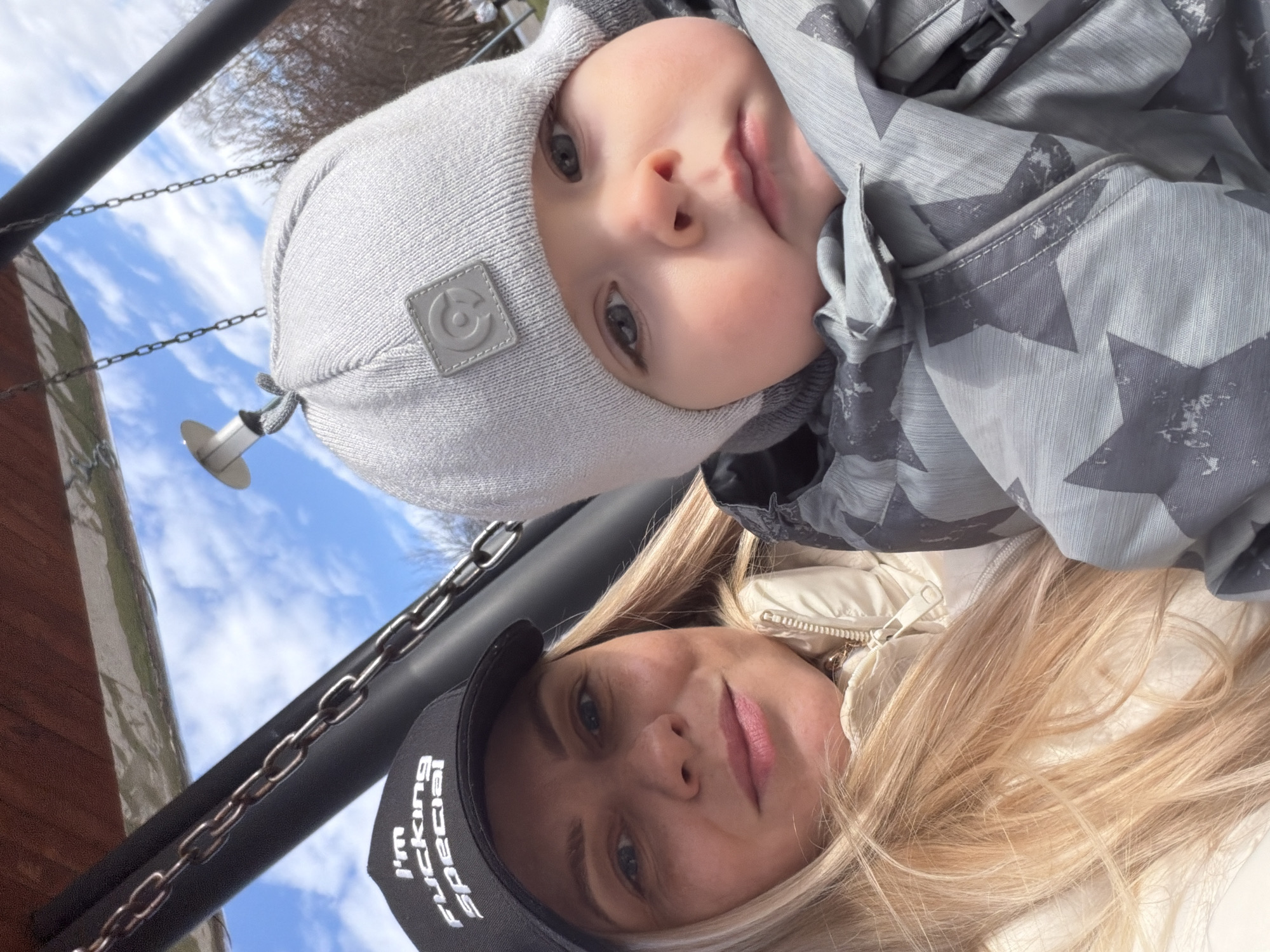 This is a close-up, rotated selfie showing a woman and a baby outdoors, likely in Votkinsk, Russia. The image is originally oriented such that the subjects appear on their sides, looking directly upwards into the camera against a bright blue sky with scattered white clouds.
The baby, appearing young, is situated in the upper portion of the frame (when viewed correctly). He wears a light grey knitted beanie hat with a small, square, grey patch featuring a circular logo (resembling a stylized 'C' or 'G'). He also has on a grey jacket with a distinct light grey star pattern. The baby has fair skin, wide blue-grey eyes, and slightly pink lips, looking with a curious or content expression towards the camera.
Below the baby is a woman, likely the mother, with long, light blonde hair. She has fair skin, light-colored eyes, and a soft, faint smile. She is wearing a light-colored (possibly cream or white) puffer jacket and a black baseball cap. The text "I'M FUCKING SPECIAL" is clearly visible in white, capitalized letters on the front of her cap.
In the background, visible against the clear sky, are dark metal chains and a dark metal bar, suggesting the subjects are on a swing set or other playground equipment. A small white cylindrical object is attached to one of the chains. Part of a blurred, reddish-brown structure, possibly wooden, is also visible. The bright lighting indicates it is daytime and sunny.
