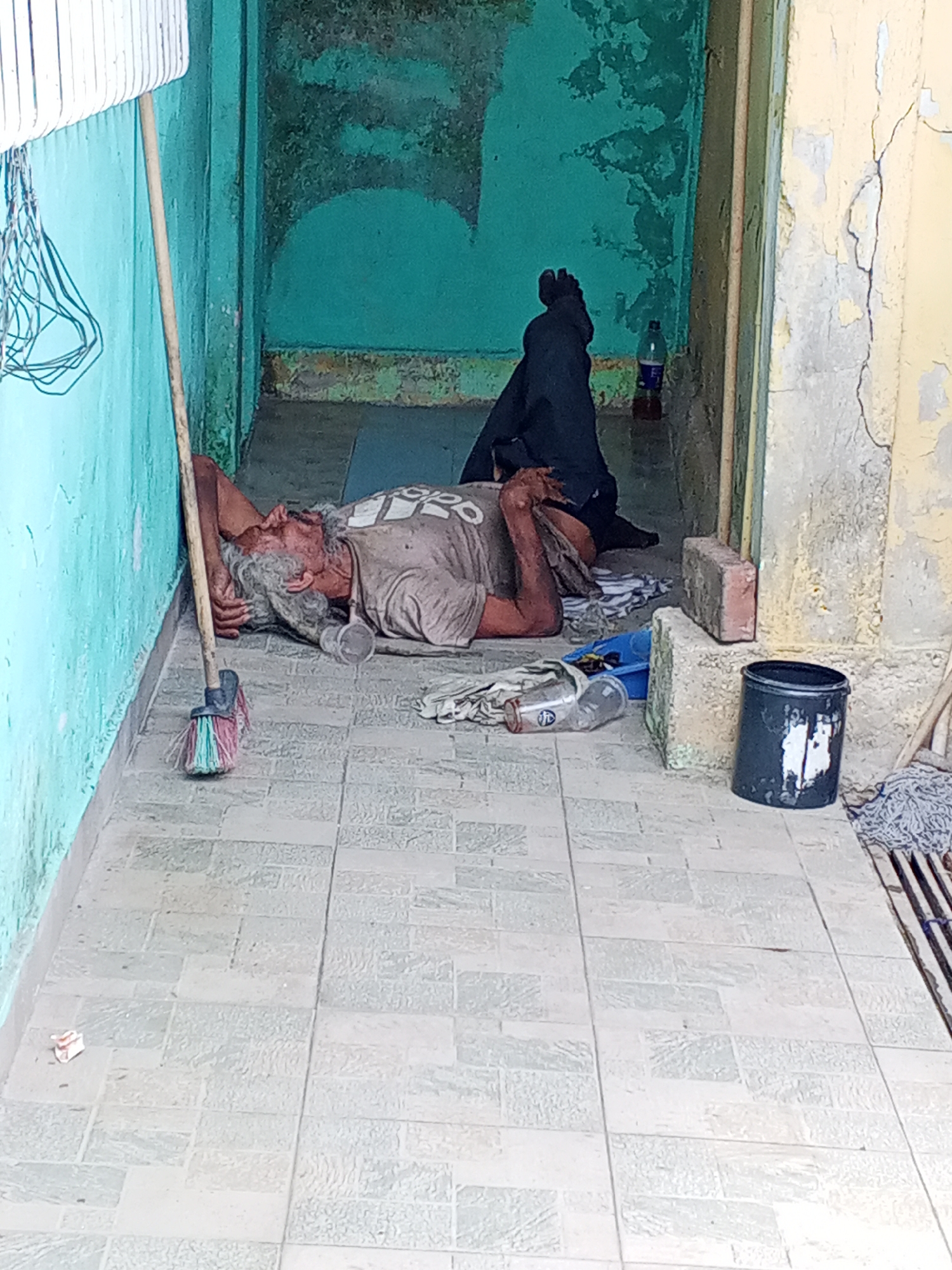 An elderly man with gray hair and a gray beard lies on a tiled floor in Tinaquillo, Venezuela. He is resting against a light blue painted wall next to a straw broom. The man is wearing a stained t-shirt. Beside him are a clear plastic cup and various empty bottles. In the background, a second person wearing dark trousers is seen lying down. A blue container and a black bucket are next to him. The environment suggests poverty and neglect. The area is a narrow outdoor corridor or passageway between two buildings with peeling paint. A mop leaning against the building to the right and a metal rack to the left are also visible.