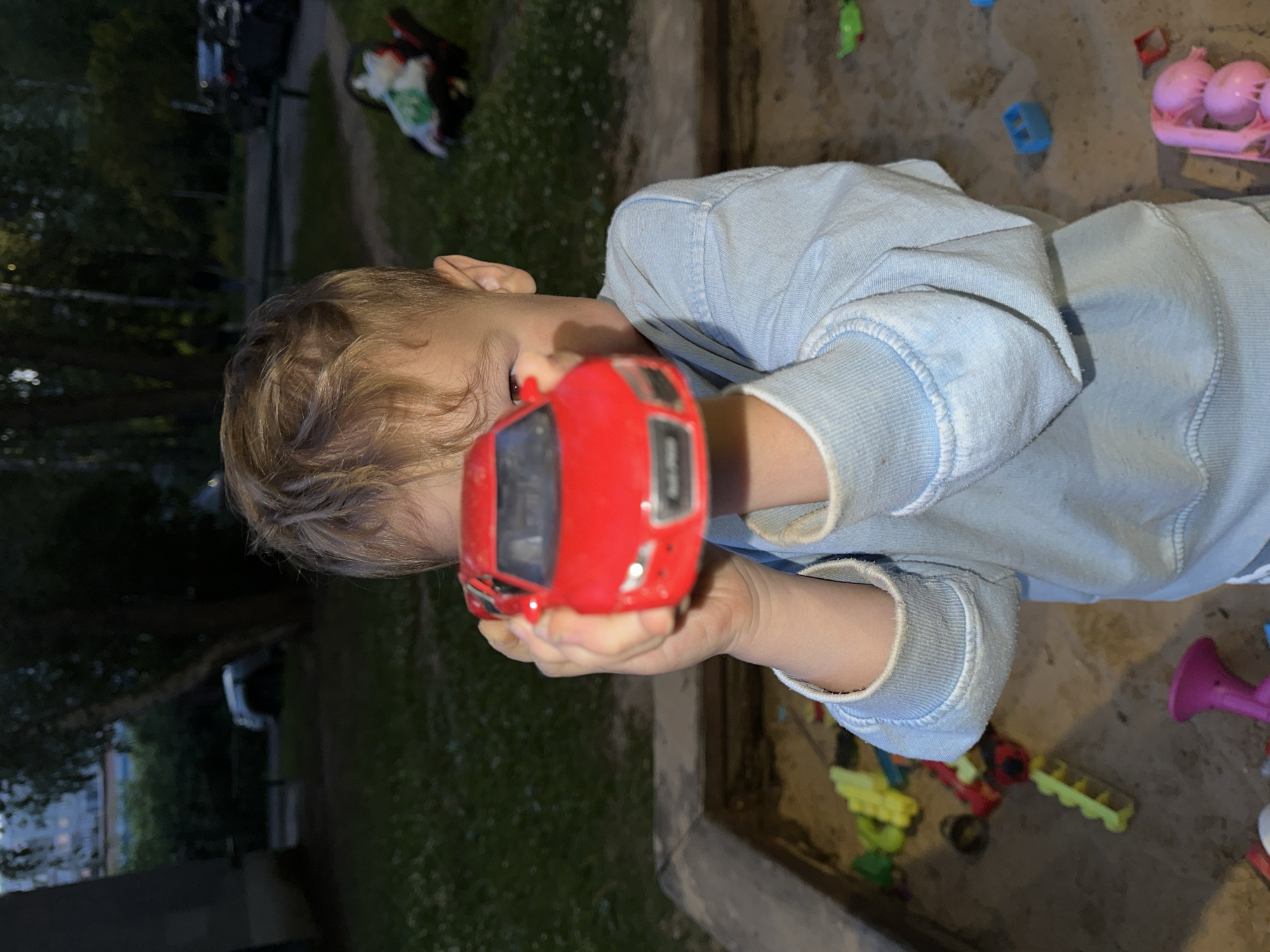 A young boy, partially visible, holds a red toy car in front of his face in a sandbox. He wears a light blue sweatshirt. The sandbox contains various plastic toys, including yellow and green pieces. In the background, grass, trees, and buildings are visible, suggesting an outdoor urban setting, possibly a playground or courtyard in Nizhny Novgorod, Russia. The lighting indicates it is dusk or nighttime.