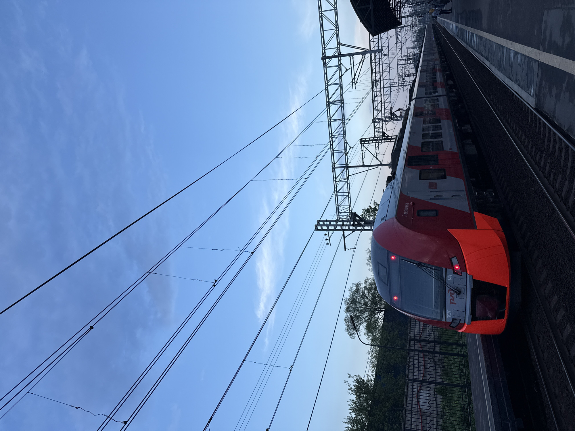 A slightly tilted, wide-angle shot captures a train on a platform in an unspecified location. The train's front is primarily red and grey. Overhead power lines extend diagonally across the left side of the frame against a blue sky with scattered clouds. A metal fence is seen behind a line of trees along the platform edge. The platform is grey and stretches into the distance, with the train parallel to its edge. Other people are partially visible toward the far end of the platform.