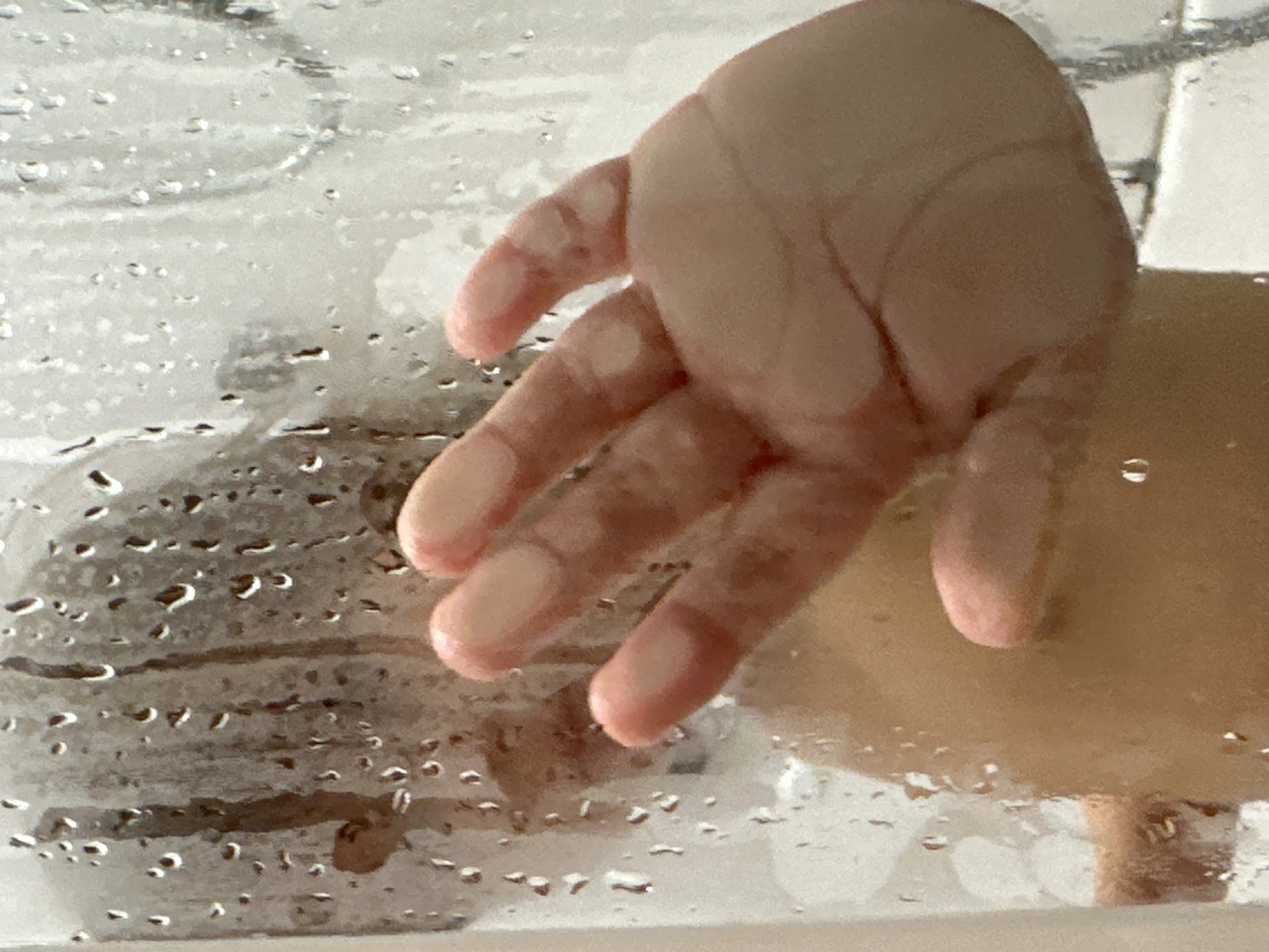 A person's hand presses against a translucent, moisture-covered surface, likely glass or plastic. Water droplets obscure the view behind the surface. Reflected or visible through the surface, there's a vague suggestion of architectural features or abstract shapes. The location is not discernible. There are no identifiable people, objects, actions, or interactions beyond the hand touching the surface.