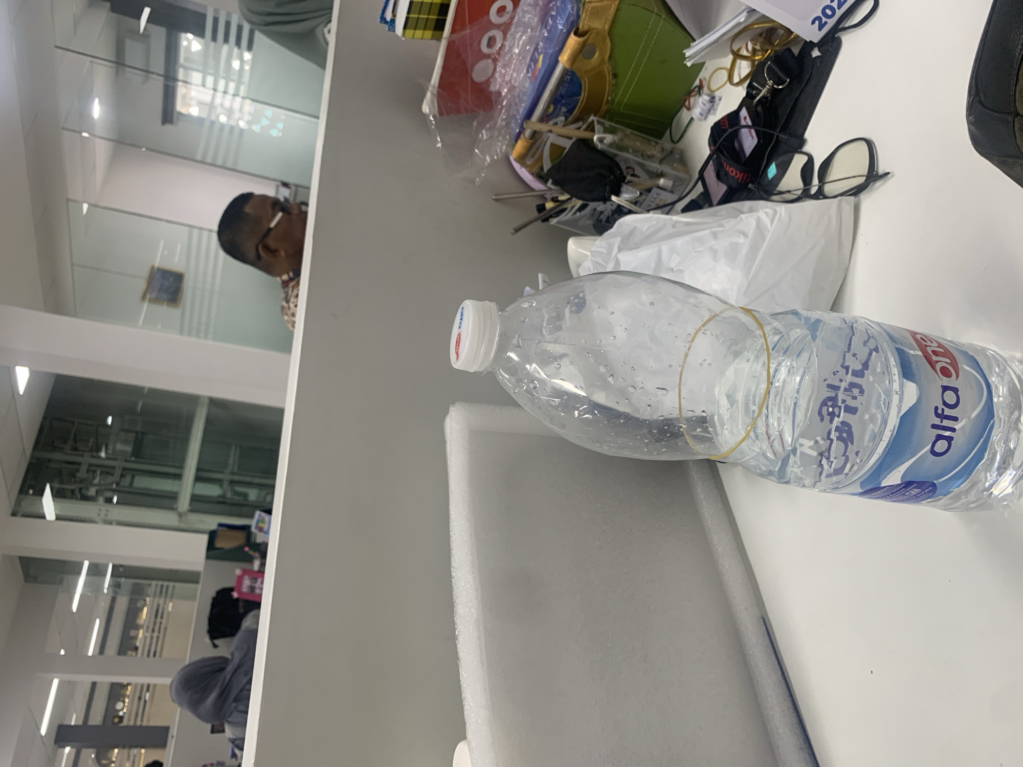 The image shows an interior view of an office environment, possibly a cubicle or open-plan workspace. In the foreground, a partially filled plastic water bottle with a yellow rubber band around it sits on a white desk. The bottle has the "alfa one" logo visible on its label. To the right of the bottle are various office supplies including a green bag, pens in a holder, a pair of glasses, and a calendar with the year "202" partially visible. In the background, through glass partitions, other office workers can be seen. One person wearing a gray hoodie is seated at a desk. Another person, a man with glasses, is also visible. The overall impression is that of a typical office setting.
