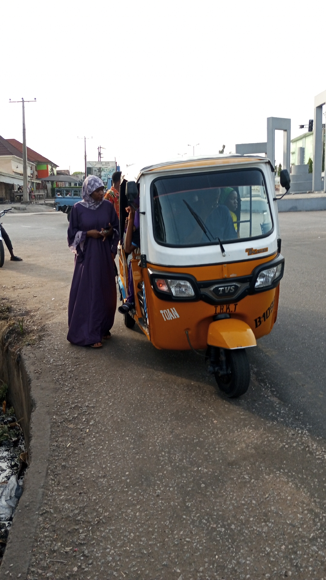 The image shows a woman wearing a purple hijab and dress standing beside an orange and white three-wheeled vehicle, likely a Bajaj or similar type of auto rickshaw. The vehicle has passengers inside. The scene is set on a paved road with a curb on the left. Background elements include buildings, a utility pole, and a structure that could be part of road infrastructure or public art. The overall impression is that this scene takes place on a road where people are getting rides or interacting with a three-wheeled transport vehicle. Given the instruction to consider the location as Adankolo, Nigeria, it can be inferred that the image captures a typical scene in a residential or commercial area of the city.