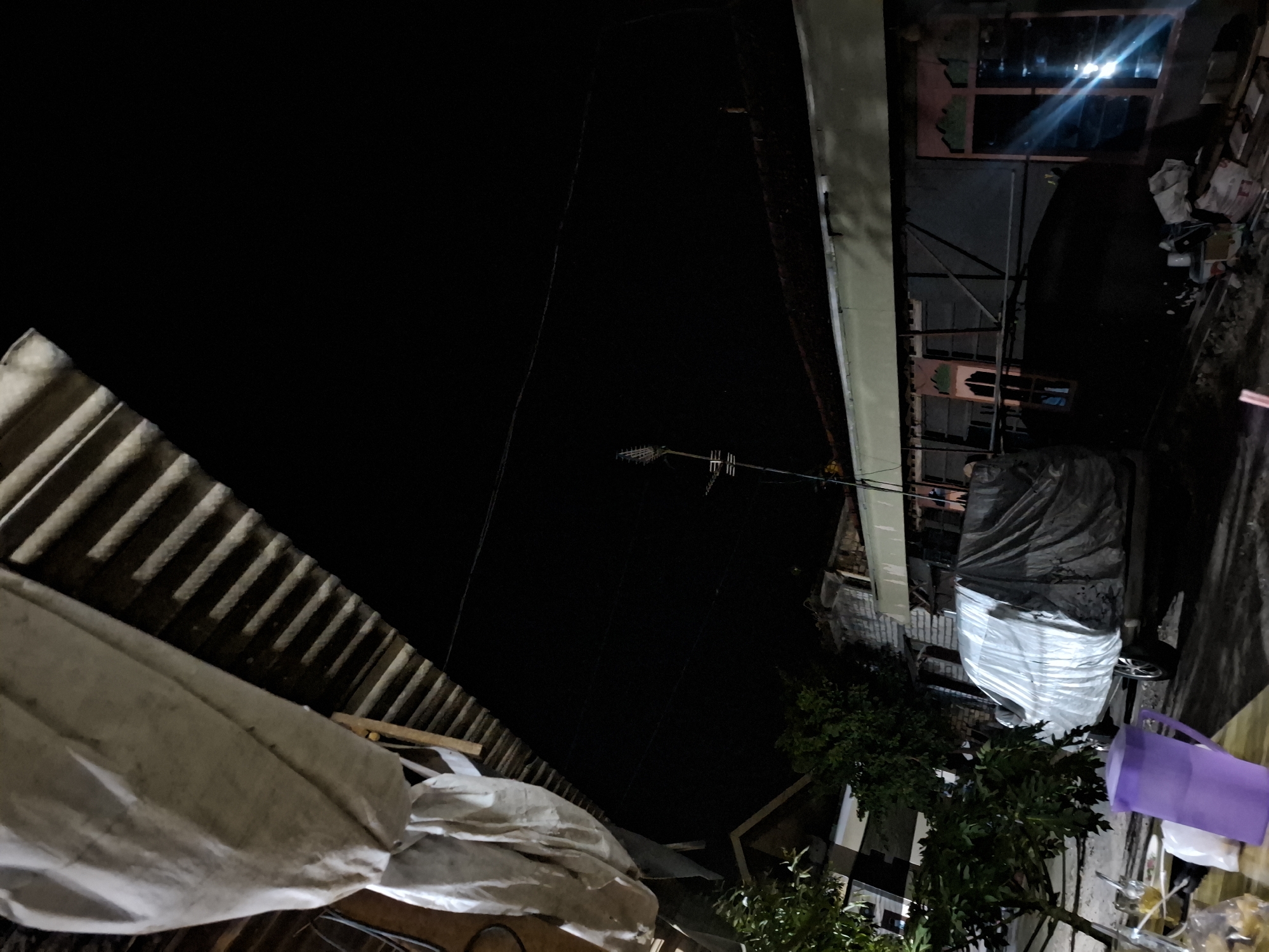 The image depicts a dimly lit outdoor scene at night in Bulubrangsi, Indonesia. In the foreground, on a wooden surface, are several objects including a purple pitcher, a glass with some liquid and ice, and plastic packaging. To the right, a vehicle is covered with a light-colored tarp. Behind the vehicle, a building with a light-colored roof and dark windows is visible. Trees with dark green foliage are present to the left of the vehicle and in the background. The sky is dark, indicating nighttime. Overhead, a corrugated metal roof structure with a light-colored fabric hanging from it is visible in the upper left corner of the image. Electrical wires are strung across the sky. A television antenna is mounted on a pole in the mid-ground.