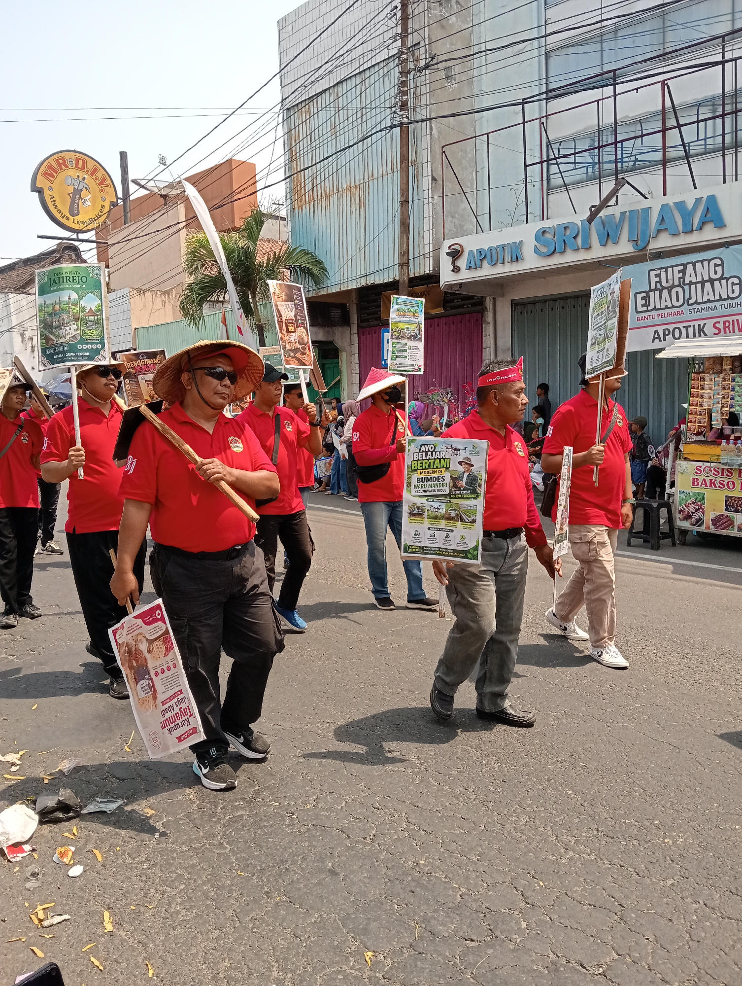This image captures a parade or procession taking place on a street in Demak, Indonesia. A group of men, most wearing red shirts and some with conical straw hats, are walking down the street, carrying various signs and what appear to be farming tools like hoes. The men are diverse in age and ethnicity. The street is paved and appears to be in a commercial area, with buildings lining the sides. One building prominently displays the sign "APOTIK SRIWIJAYA." Another building has a circular sign with "MR.D.I.Y." The weather appears to be sunny and clear. The overall atmosphere suggests a local event or celebration.