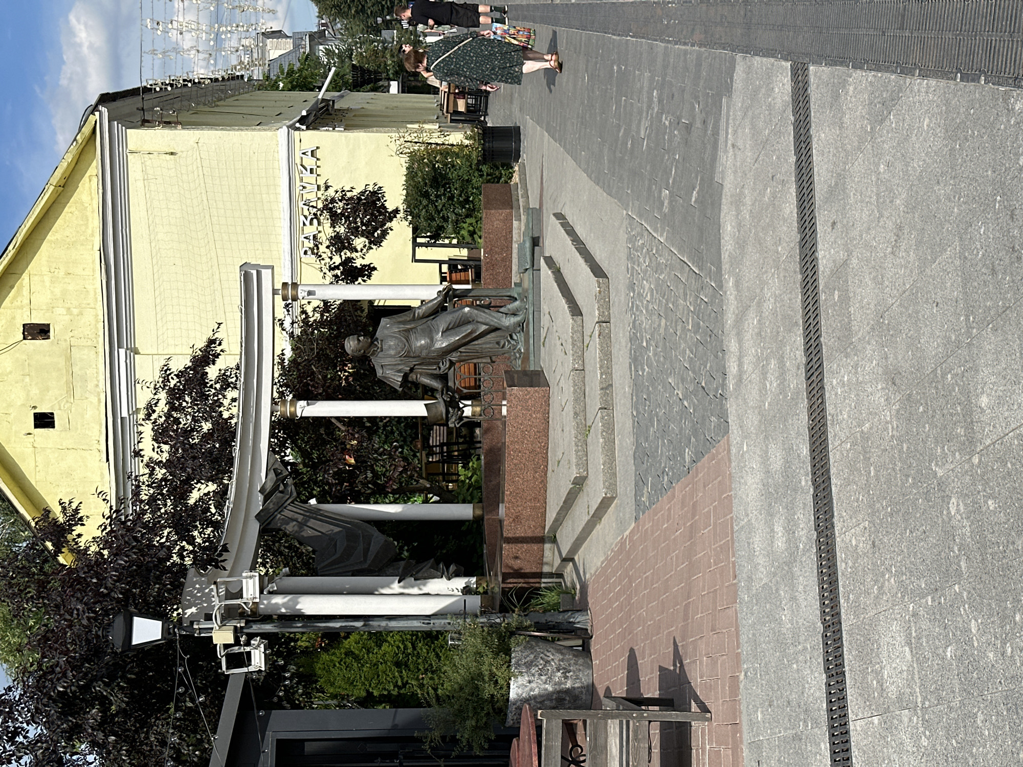 A full shot captures a sunny day in what appears to be a public square or pedestrian area in Putyatino (Semivragovskaya S/A), Russia. A statue of a man in academic attire, leaning on a pedestal, is a central focus. Behind the statue, a light yellow building with a decorative cornice stands, featuring a sign with Cyrillic lettering. The ground is paved with cobblestones and granite slabs, with a prominent grate running across the foreground. To the right, two women walk away from the camera, one carrying a shopping bag. To the left, outdoor seating and planters are visible, suggesting a cafe or restaurant setting. Lush green trees and a lamppost add to the urban landscape.