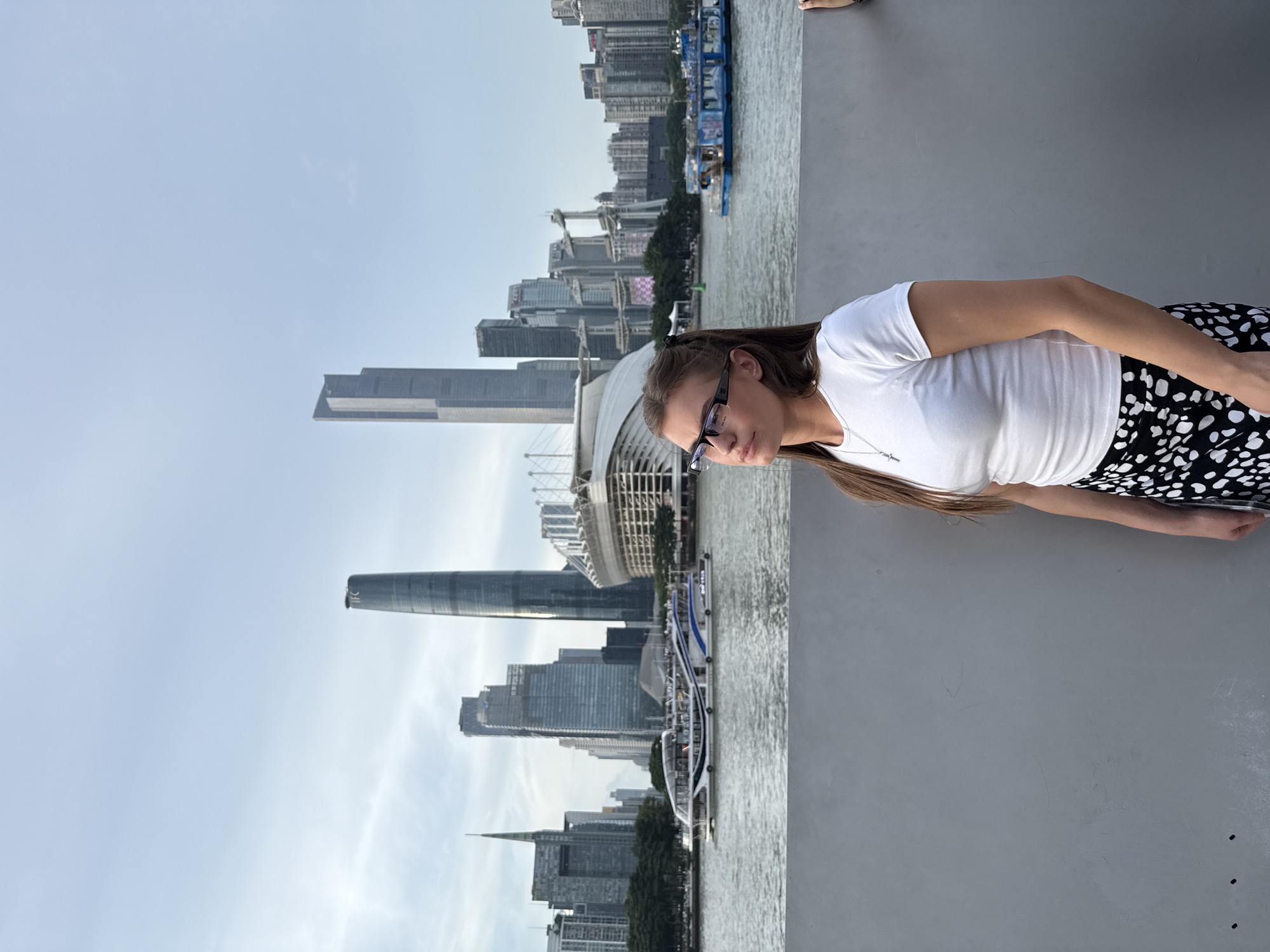 A young woman with long brown hair, wearing glasses, a white t-shirt, and a black and white patterned skirt, stands in the foreground looking upwards and to the right. She is positioned in front of a grey wall. Behind her, a wide river flows, with several boats on its surface. In the background, a modern cityscape unfolds, featuring numerous skyscrapers of varying heights and architectural styles, along with a large stadium-like structure. The sky is overcast. The location appears to be the Pearl River waterfront in Yuexiu District, China, likely in the city of Guangzhou, given the prominent skyscrapers like the Canton Tower and the Guangzhou IFC.