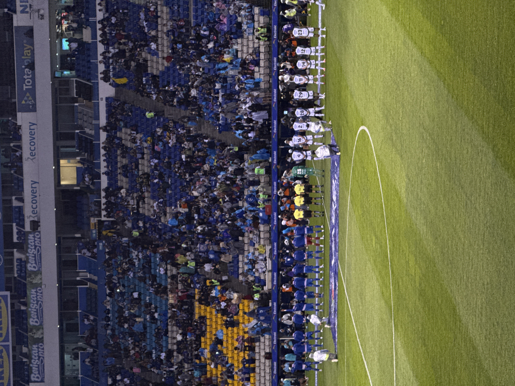 The image shows a soccer match at Estadio Cuauhtémoc in Puebla, Mexico. A crowd fills the stadium. Players in white and blue uniforms stand on the grass field. A row of players in dark blue uniforms stands beside them. Signage for various sponsors, including "Electrolit", "Recovery", and "Tota play", is visible around the perimeter of the field and stadium.