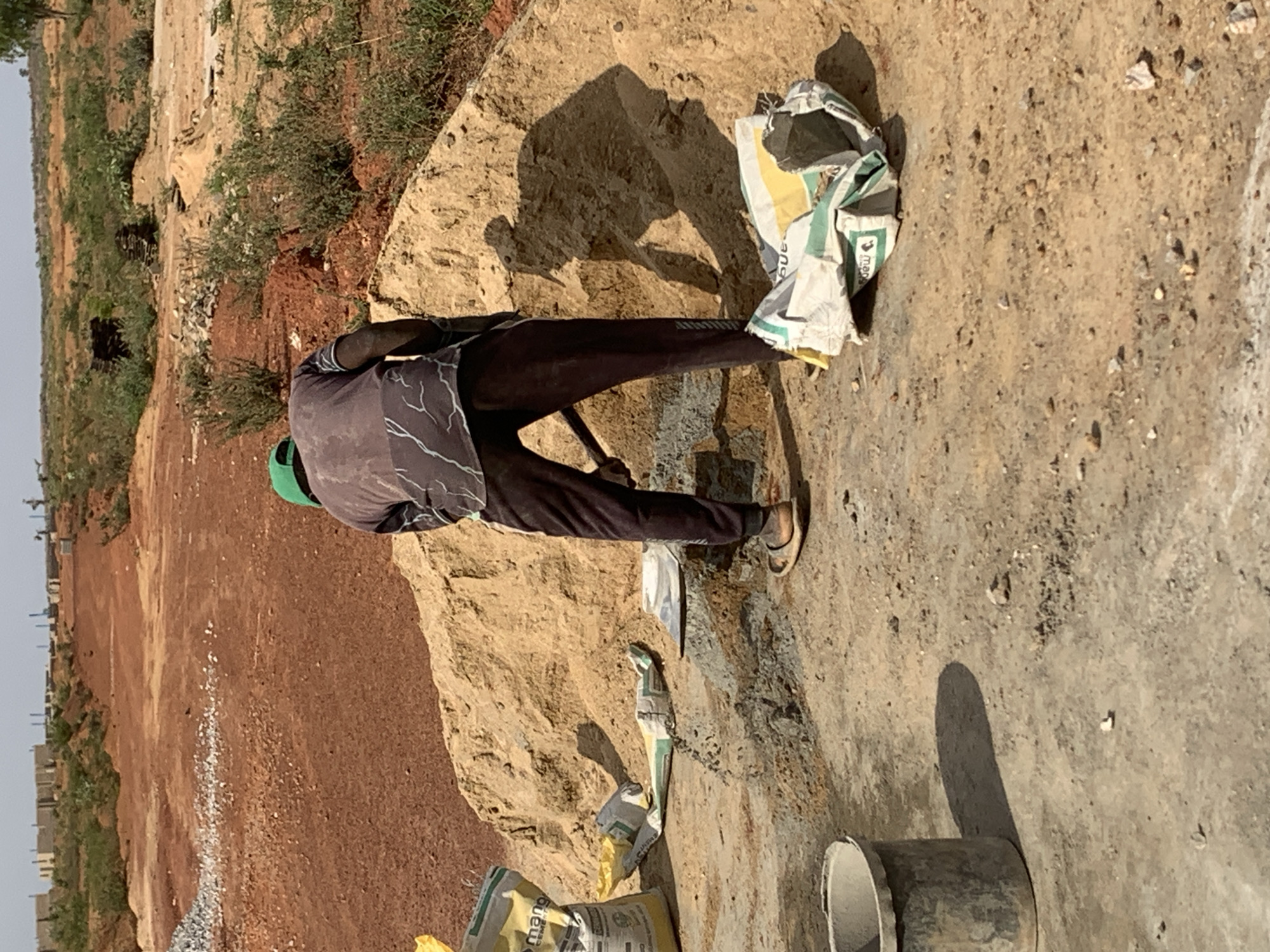 A man wearing a green head covering, a grey t-shirt with dark patterns, and dark pants is standing outside at a construction site in Kujama, Nigeria. He is near a pile of dirt/earth/sand, some open cement bags, and a piece of construction equipment. The background shows buildings and vegetation.