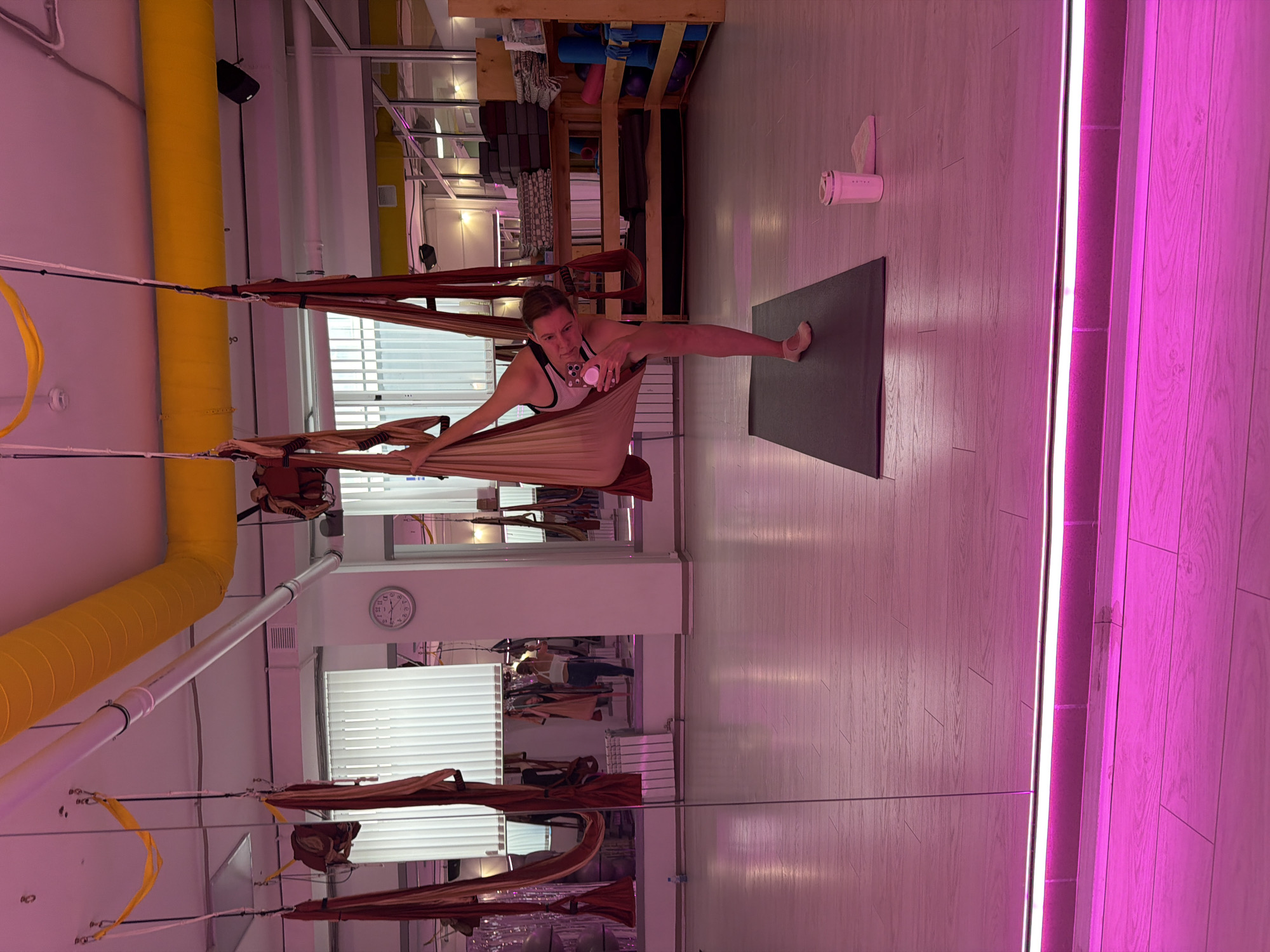 A woman is performing aerial yoga in a studio. She is suspended in a fabric hammock, with her legs extended and one arm reaching up. She is holding a pink phone in her other hand, appearing to take a selfie. The studio has a wooden floor and is lit with pink and yellow lighting. There are several other fabric hammocks hanging from the ceiling, as well as yoga mats and other fitness equipment visible in the background. The image does not provide information about the city of Chaikovsky, Russia.
