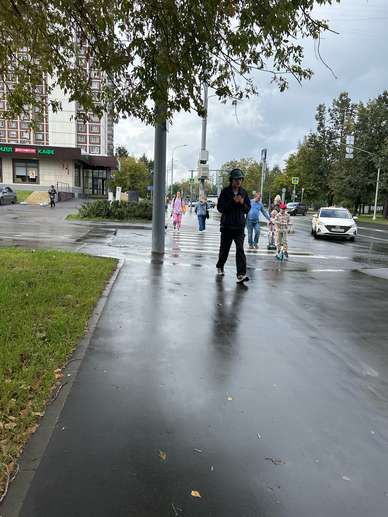 On a cloudy day in Moscow, Russia, people cross a wet street at a pedestrian crossing. The road reflects the overcast sky and surrounding environment. On the left, a sidewalk with green grass leads to a building with a cafe. On the right, trees line the street. In the foreground, a man in a dark jacket and pants walks across the street. Behind him, a woman with a dog on a leash crosses the street, followed by a family with two children on scooters. A white car, possibly a taxi, drives on the right side of the road. The image captures a typical urban scene in Moscow, with everyday activities taking place amidst the city's architecture and weather.