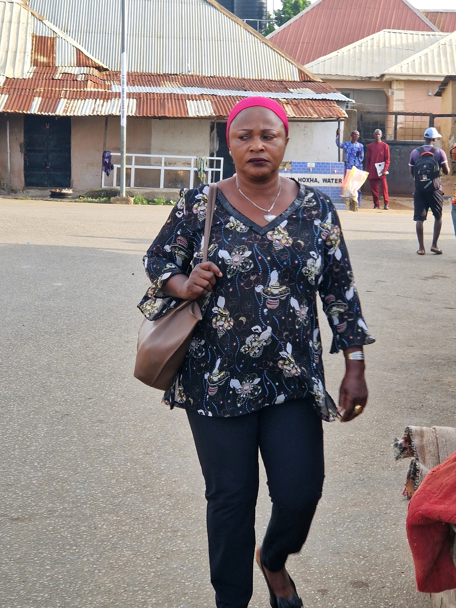 In the city of Kabong, Nigeria, a Black woman with a pink headscarf and dark lipstick walks across a paved surface. She is wearing a dark, floral-patterned shirt and black pants, carrying a brown shoulder bag. Behind her, two men stand near a blue structure with white lettering that reads "HOXHA, WATER." Further in the background, another man in a red outfit and a person wearing a backpack are visible. The environment appears to be an outdoor, semi-urban area with corrugated metal roofs on buildings.