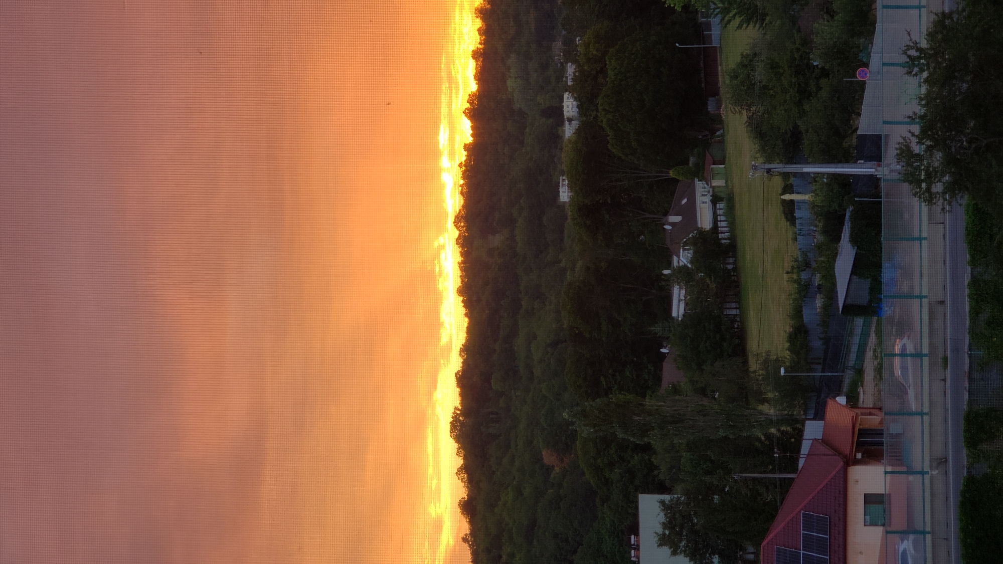 The image depicts a sunset view from an elevated position, likely a residential building in Prešov, Slovakia. The sky is dominated by a gradient of orange and pink hues, transitioning from a vibrant yellow glow at the horizon to a muted rose color higher up. Below the sky, a dense, dark green forest covers a rolling hill. The treeline is silhouetted against the bright sky, with glimpses of scattered houses nestled within the greenery. In the foreground, a red-roofed house with solar panels is visible, along with a fence and a road with cars. A streetlamp and some smaller trees are also present. The overall impression is one of a tranquil evening in a suburban or semi-rural area of Prešov.