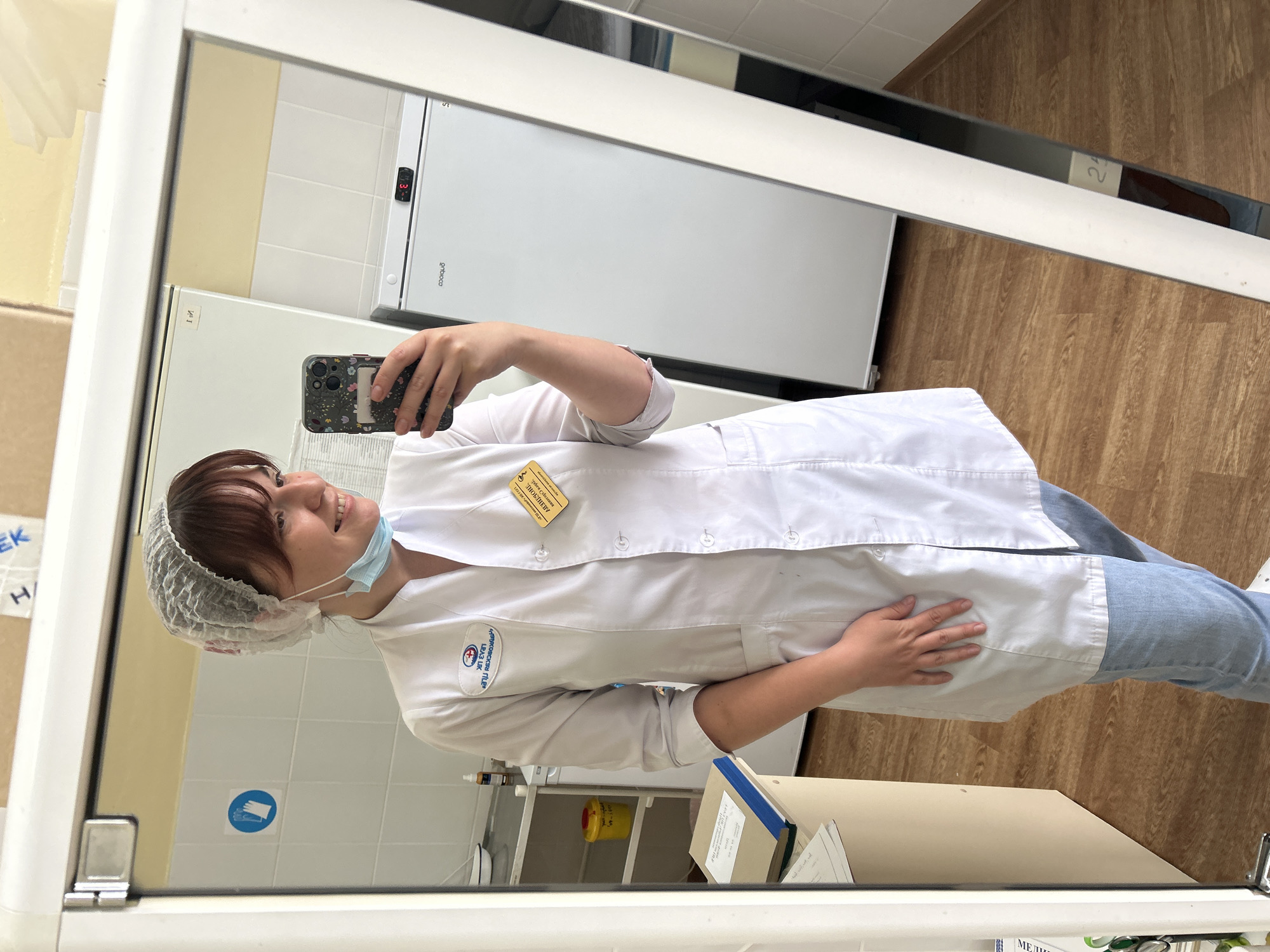 A woman wearing a white lab coat, a blue surgical mask, and a white hairnet is taking a selfie in a mirror. She is standing in what appears to be a lab or medical setting. A white refrigerator unit is visible behind her. A yellow name tag is pinned to her coat, and the lab coat has a logo on the left arm. The background reveals white tiled walls, a shelf with various items, and wooden flooring. The city of Фоки, Russia is not verifiable in this image.