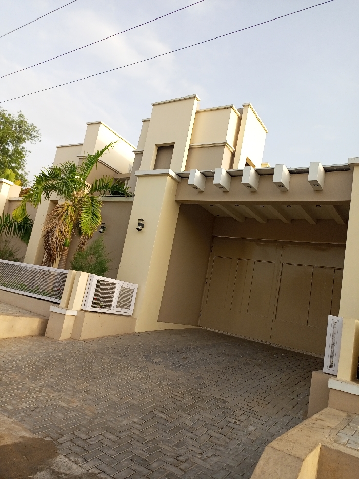The image shows a large, beige-colored, modern house with a carport in Maiduguri, Nigeria (latitude: 11.82, longitude: 13.154). The house has a white awning above the carport entrance, and a large, double-door garage door. The garage door is closed and made of tan-colored metal. There is a palm tree in front of the house, and a small, white ventilation grate on the side of the house. The yard is covered in gray cobblestones. The sky is a light blue and there are power lines running above the house. There are no people visible in the image.