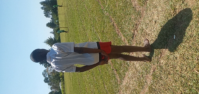 The image shows a young boy standing on a grassy field in Burundu, Kenya, at coordinates 0.53 latitude and 34.746 longitude. The boy is wearing a white shirt and red shorts. He has his hands clasped behind him, and he is looking at two other people in the distance. The field is covered in short green grass. Two other figures are visible in the distance, moving away from the boy. The sky is clear and blue. The image appears to be taken on a sunny day. The trees in the background are mostly deciduous. The field is relatively flat and without noticeable obstructions. The scene is peaceful and serene.