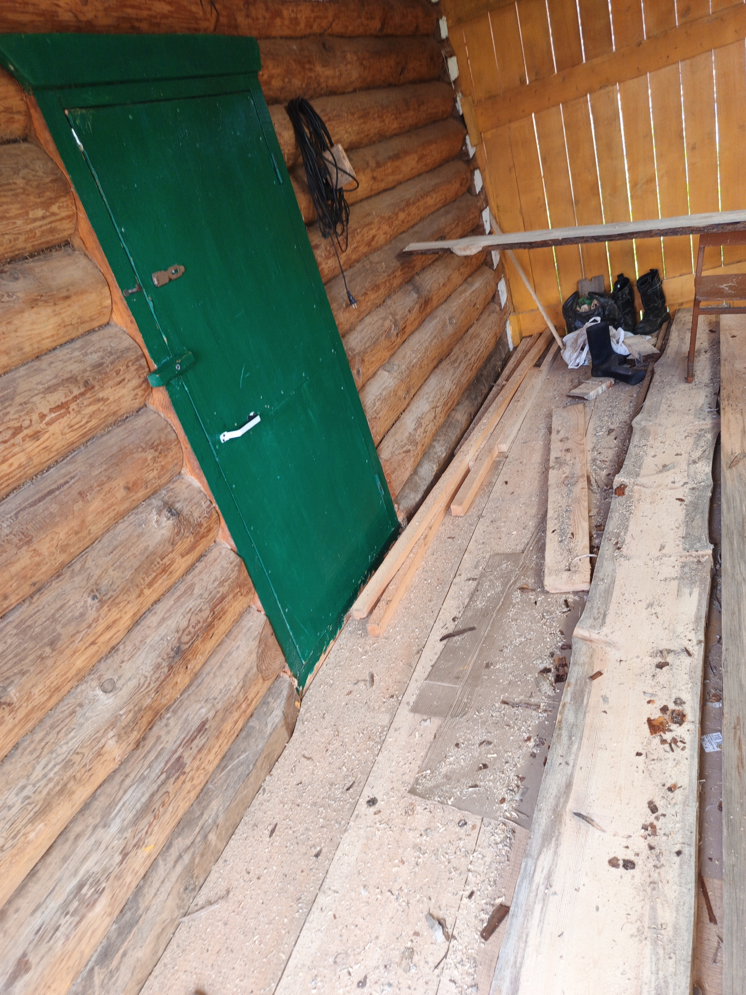 The image depicts the interior of a rustic structure, likely a shed or storage area, constructed from rough-hewn logs. A prominent, emerald-green door is visible on the left, angled with the slope of the building. The door has a simple latch and a white handle. To the right of the door, a coiled black cable with a white electrical plug hangs from the wall.
The floor of the structure is made of wooden planks, covered in sawdust and wood shavings, suggesting recent carpentry or construction work. Various wooden planks, some rough-cut and others appearing to be reclaimed lumber, are scattered across the floor.
In the background, a wooden shelf made of a single plank is supported by metal legs. Underneath this shelf, a black plastic bag filled with debris is present, along with a pair of black rubber boots and another boot lying on its side. To the far right, a simple wooden chair with a metal frame is visible, partially obscured by a wooden plank. The walls and ceiling are also constructed from wooden planks, with a warm, yellowish hue. The overall impression is one of a working space or storage area that is in a state of disarray. The location is identified as Islambaevo, Russia.
