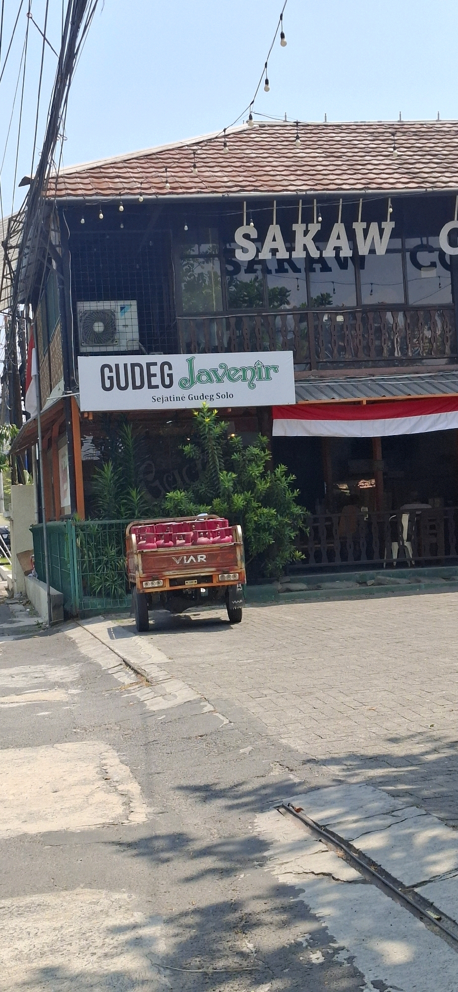 In Colomadu, Indonesia, a three-wheeled truck is parked on a paved area in front of a building. The truck is red and its bed is filled with pink gas cylinders. The building has a tiled roof and a dark facade with windows. A sign above the windows reads "SAKAW COFFEE". Another sign below, on a white banner, reads "GUDEG Javenir" with the subtitle "Sejatine Gudeg Solo". An air conditioning unit is visible mounted on the exterior wall of the building. The scene is illuminated by bright sunlight, casting shadows on the ground.