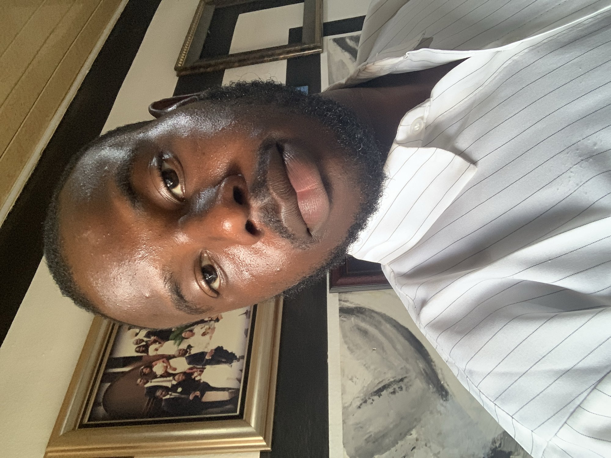 A close-up selfie of a Black man with a beard, wearing a white and gray striped shirt. He is indoors. Behind him are picture frames on a wall. No evidence suggests this image was taken in Jos, Nigeria.