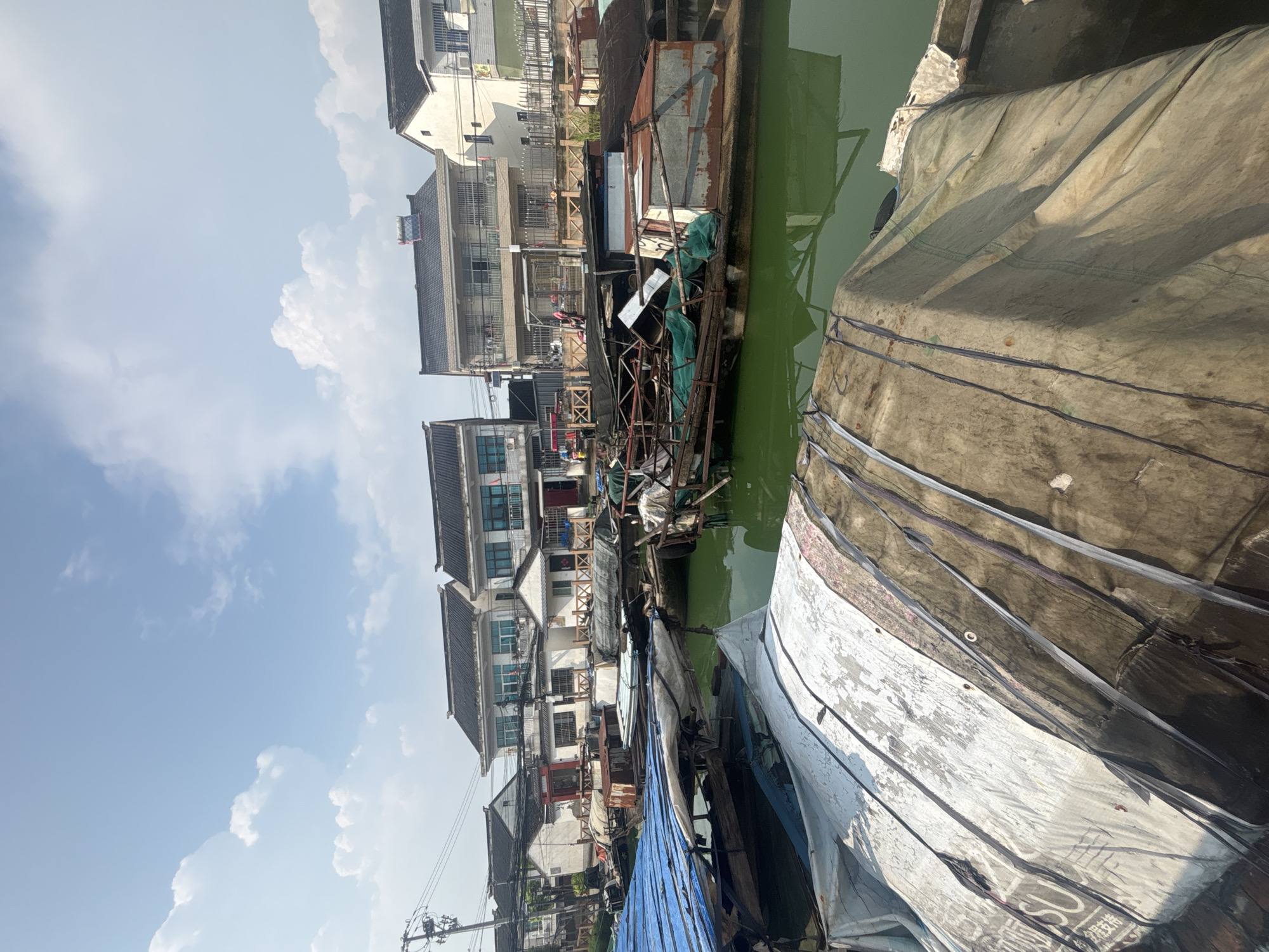 The image depicts a canal scene in Wuzhong District, China, likely in a traditional water town. Several boats are moored along the canal, some appearing weathered and covered with tarps and various materials. The water is a murky green color. In the background, traditional Chinese houses with dark tiled roofs and white walls line the canal. The sky is blue with scattered white clouds. Electrical wires and poles are visible along the houses. The overall impression is of a working canal with residential structures. There are no people clearly visible in the scene.