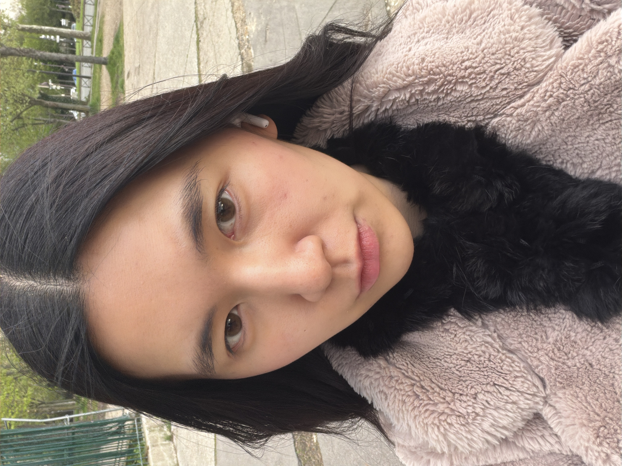 A medium close-up portrait shows a young woman with straight black hair and light brown eyes. She is wearing a light brown, fluffy coat with a black fur collar. A white earbud is visible in her left ear. She is looking directly at the camera with a slight smile. The background shows trees and a metal railing, suggesting a park setting. Based on the style of the railing and the trees, the location is likely in a park in Paris, France. The ground visible in the background appears to be paved. The lighting suggests it is daytime.