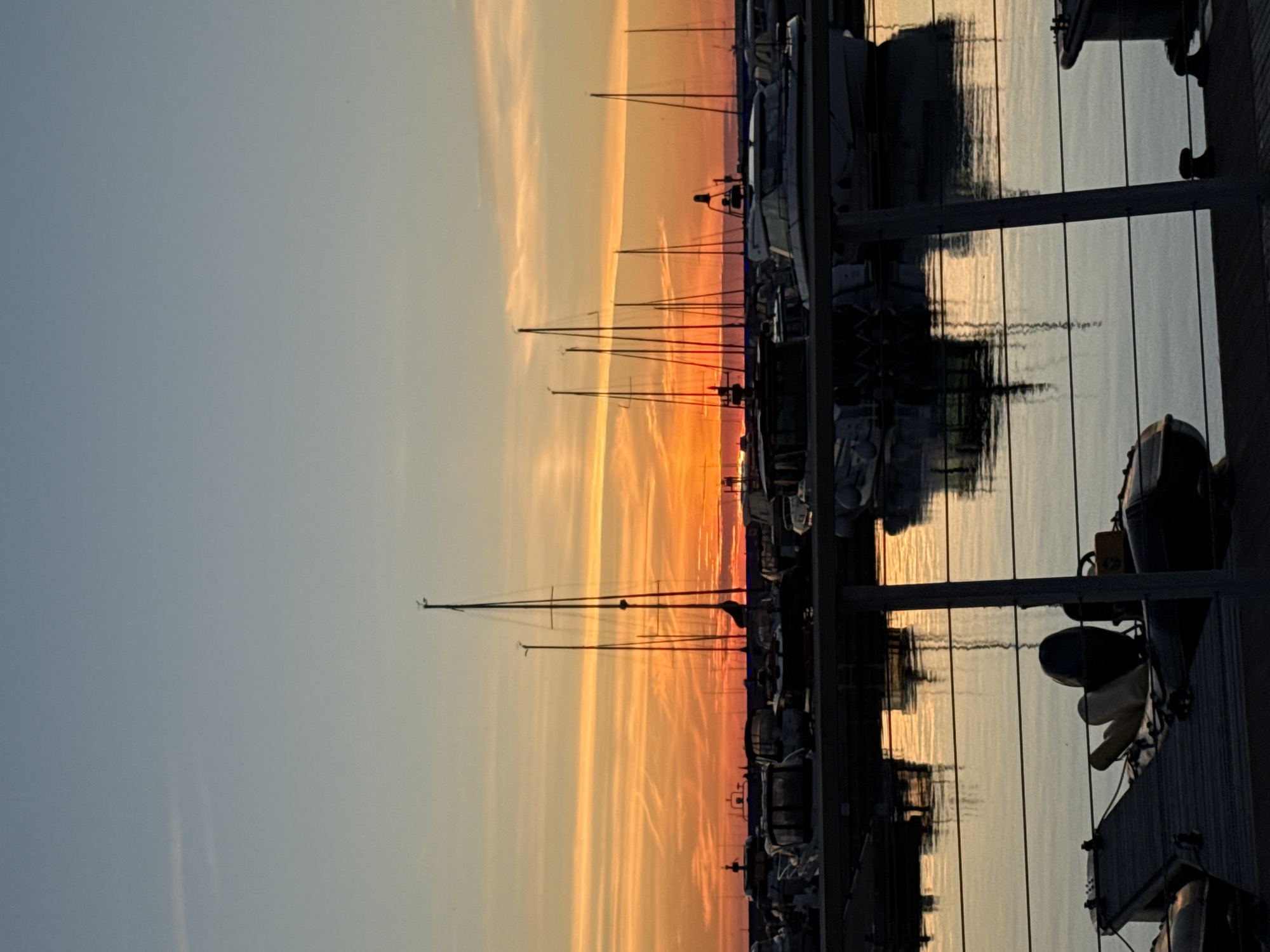 The image depicts a harbor scene at sunset in Tallinn, Estonia. The sky is a gradient of blue and orange, with streaks of clouds reflecting the vibrant sunset colors. Several sailboats with tall masts are docked in the harbor, their silhouettes stark against the colorful sky. In the foreground, various motorboats and smaller vessels are moored. The water is calm, reflecting the boats and the sky. The scene appears to be from a waterfront promenade or dock, with railings visible in the foreground. The overall impression is one of tranquility and natural beauty as day transitions into evening.