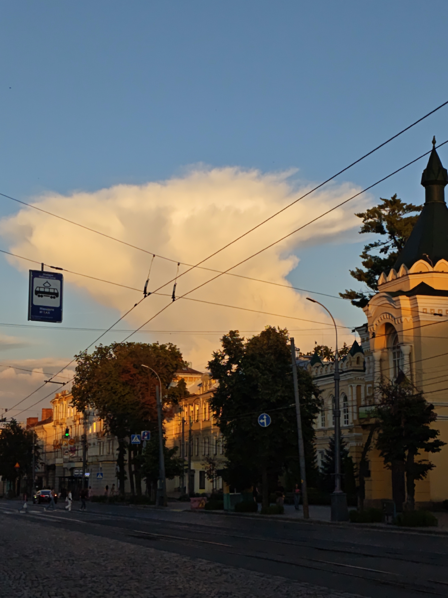 The image captures a street scene in Vinnytsia, Ukraine, during what appears to be late afternoon or early evening, given the warm, golden light illuminating the buildings and sky. The sky is a vibrant blue with a large, dramatic cumulus cloud formation glowing with a peach hue, possibly indicating sunset. Overhead, electrical wires crisscross the frame, supported by poles.
On the left side of the image, a blue and white sign indicating a tram stop is suspended from wires, displaying a tram icon and text that appears to be in Ukrainian, reading "Трамвай № 1, 4, 5" (Tram No. 1, 4, 5).
The street itself is paved with cobblestones in the foreground, and further down, there are tram tracks. Several pedestrians are visible crossing the street at a pedestrian crossing. A dark-colored car is also present in the street.
Buildings line the street, with architecture that suggests a historical or classical style, featuring ornate details and arched windows. One prominent building on the right has a distinctive dark green spire and a yellow facade. Trees with green foliage are scattered along the sidewalk and in front of the buildings.
The overall atmosphere is that of a functional urban environment bathed in the soft, warm light of a setting sun, creating a picturesque scene.