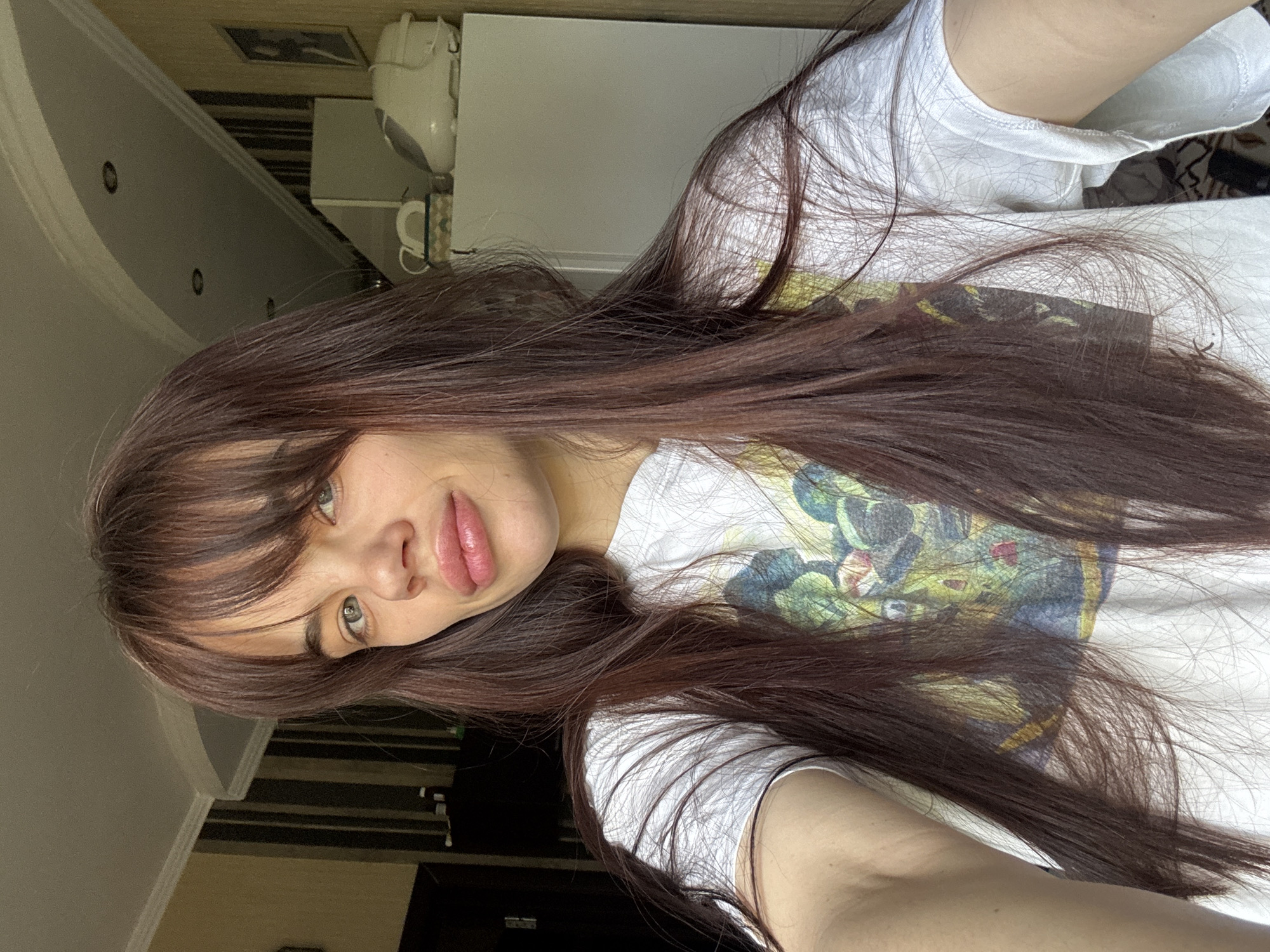 A person is taking a selfie in an indoor environment, possibly a home. The individual has long, dark brown hair with bangs and is wearing a white t-shirt with a colorful graphic design. The background includes a glimpse of a room with a wall and what appears to be a kitchen appliance. No specific landmarks or details definitively indicate the location as Rostov-on-Don, Russia.