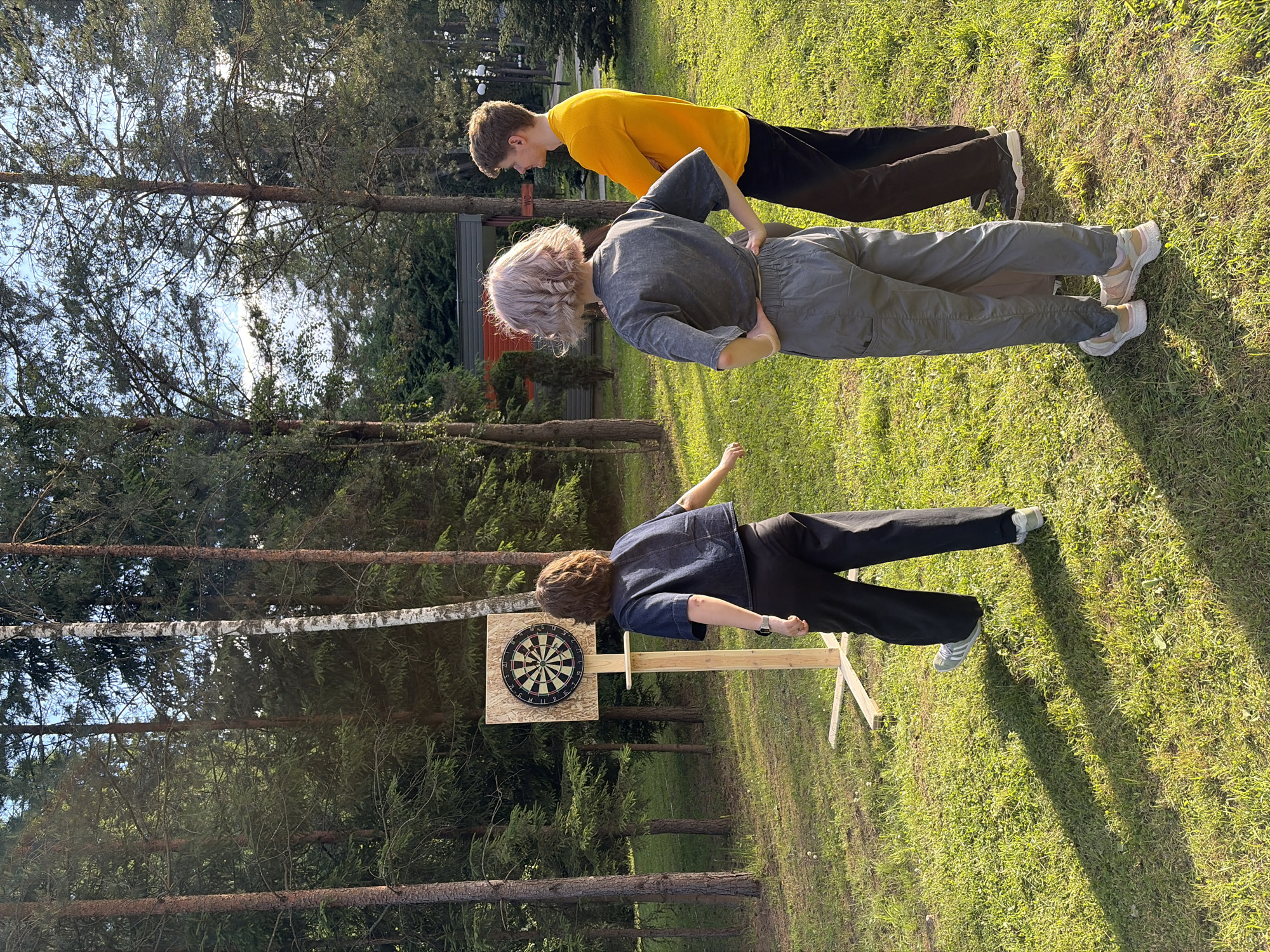 In the Zhdanovichi rural council, Belarus, three individuals are gathered on a grassy area surrounded by trees. A dartboard is mounted on a wooden stand, and one person is facing it, seemingly mid-throw. Another individual stands to the side, observing, with their hands on their hips, and a third person stands behind them, looking in the same direction. The setting appears to be outdoors, possibly a park or recreational area, with a building visible in the background. The lighting suggests it is daytime, with sunlight casting shadows on the grass.