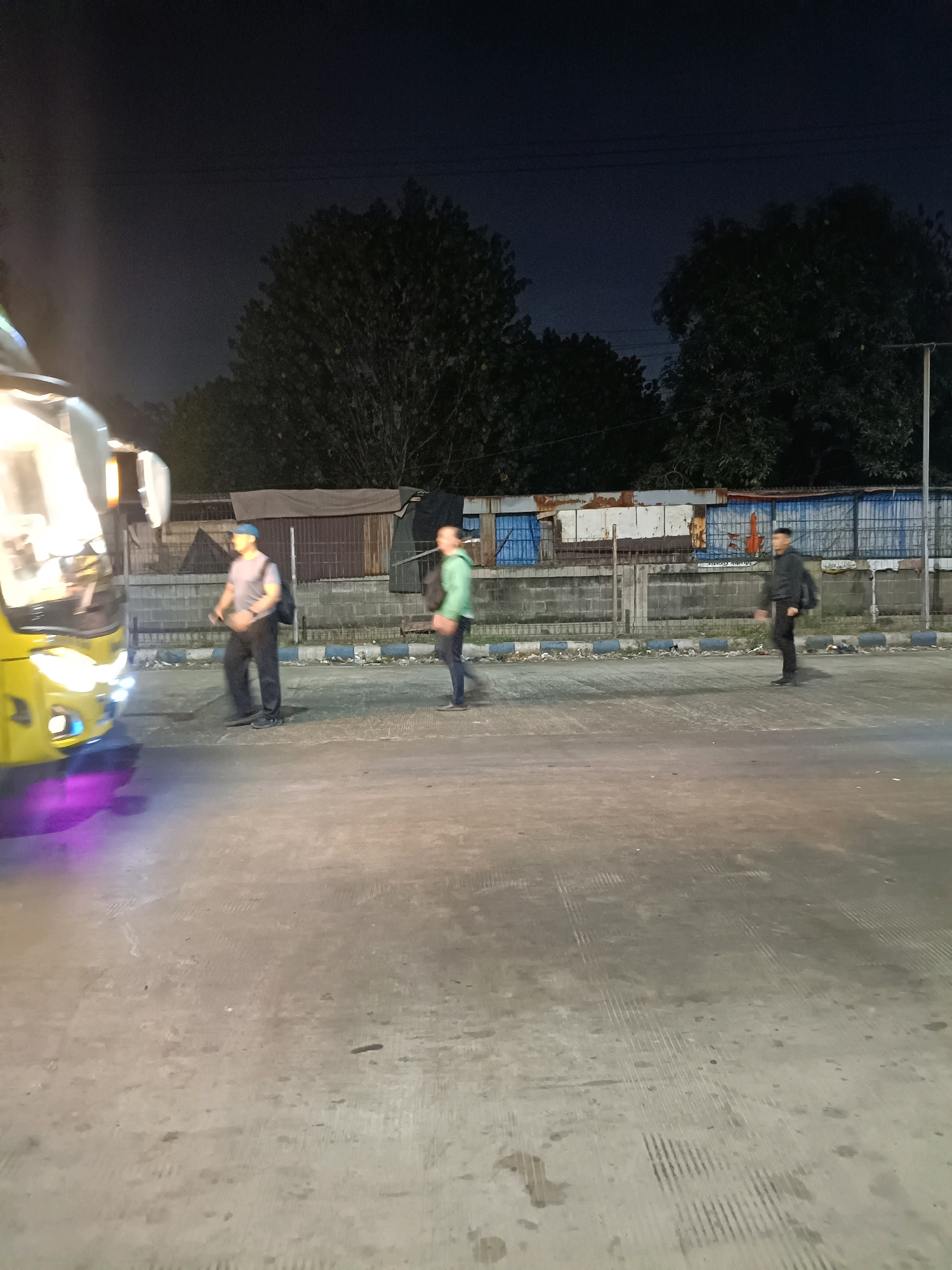 The image depicts a nighttime scene on a paved road in Kedungrejo, Indonesia. In the foreground, a yellow vehicle, possibly a bus or van, is partially visible on the left side, with its headlights illuminated and casting a purple glow on the ground. Three individuals are present on the roadside. The person closest to the vehicle is wearing a grey t-shirt, black pants, a blue cap, and a backpack. To their right, another person in a green hooded jacket and dark pants is walking away from the camera. Further to the right, a third person in a black jacket and black pants is walking towards the right side of the frame. Behind the individuals, there is a fence and a row of buildings or structures with corrugated metal and various colored panels. Trees with dark foliage are visible in the background against a dark night sky. The road surface appears somewhat rough and shows signs of wear.