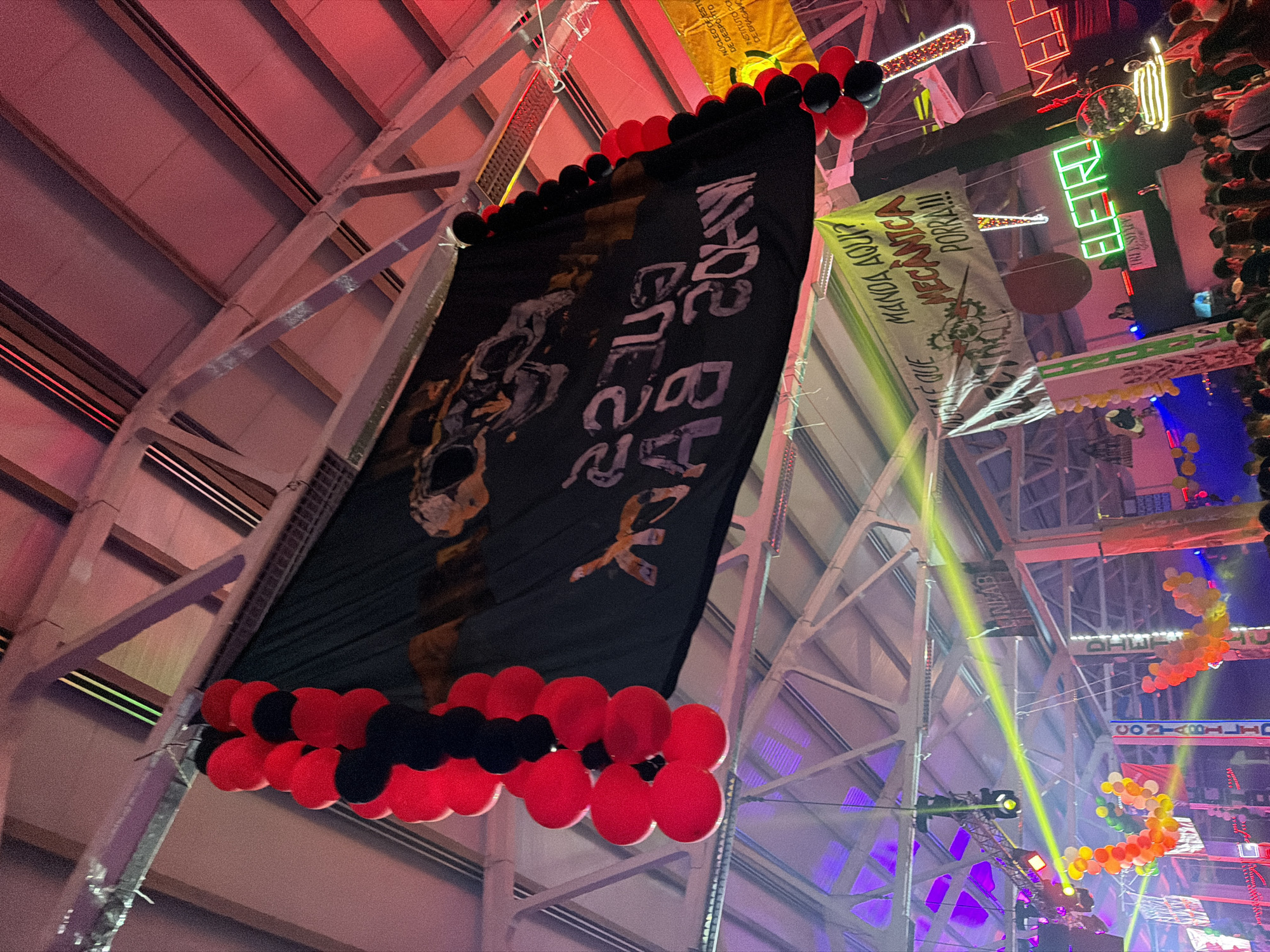 This image captures an indoor nighttime event, likely a university or student gathering, held within a large hall featuring an industrial-style ceiling with prominent metal trusses and beams. The space is vibrantly decorated with numerous banners, balloons, and colorful lighting.
A large black banner hangs centrally from the ceiling, adorned with clusters of red and black balloons at its top and bottom. This banner displays a graphic that appears to be a stylized skull or head, along with inverted white text, partially legible as "MADE BY 2024".
Around the venue, several other banners are visible, suggesting different student groups or academic departments. A yellow banner reads "NÚCLEO D DE DESPO INSTITUTO DE BRAGH," directly referencing the Instituto Politécnico de Bragança. Another light green banner proclaims "QUEM É QUE MANDA AQUI? MECÂNICA PORRA!!!" with a gear-like logo, indicating a mechanical engineering student association. Further in the background, banners with "CONTABILID" (likely Accounting) and "LINEAB" are discernible.
The atmosphere is festive, illuminated by dynamic lighting, including red, purple, yellow, and green spotlights casting beams across the space. Neon signs spell out "ELETRO" and "ELETROMED". On the far right, a crowd of people can be seen, possibly observing the event from an elevated vantage point, underscoring the social and celebratory nature of the occasion. Red, black, yellow, orange, and white balloons are arranged throughout, with some forming an arc shape in the background. The event appears to be a lively student party or festival in Bragança, Portugal.