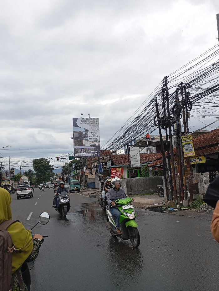 The image shows a street in Katapang, Indonesia (latitude: -6.998, longitude: 107.554). The road is wet from recent rainfall, and there are puddles in the road. There are several motorbikes traveling on the street. One motorbike is being driven by a man wearing a helmet. The man is wearing a long-sleeved shirt and jeans. There are several people in the background, including a person on a scooter who is wearing a helmet and another person in a car. The road is surrounded by buildings and trees. There are many electrical wires running overhead. A large billboard is visible on the right side of the image. The billboard advertises a company that specializes in industrial goods and services. There are several signs on the buildings in the background. The signs are in Indonesian.