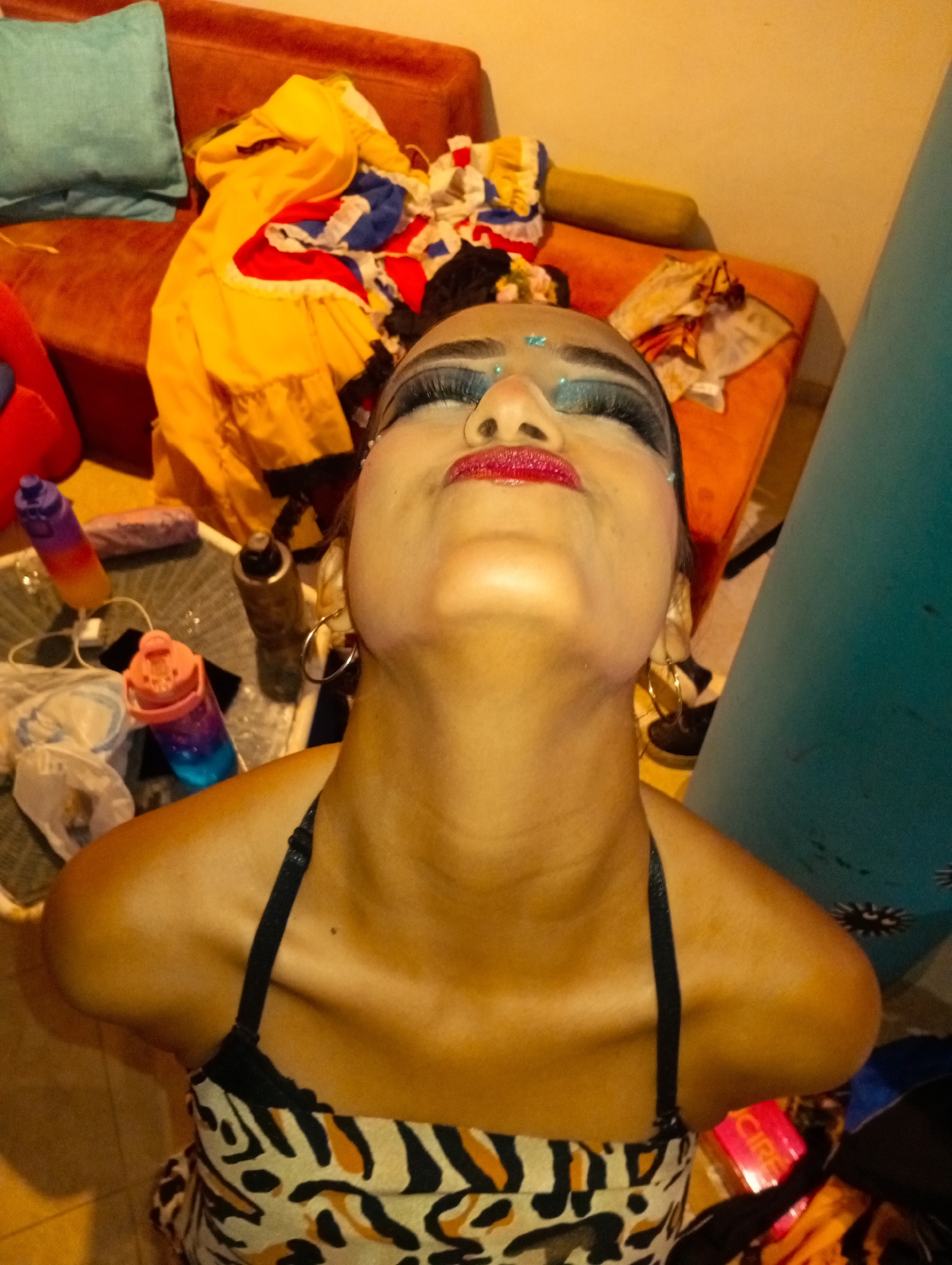 A woman with heavy makeup and wearing a black and orange halter top is photographed from a low angle, looking upward. Her makeup includes red lipstick, heavy eyeliner, glittery eyeshadow, and glued-on gems. She is wearing hoop earrings. In the background, there is a messy area with clothing draped over a couch and a table cluttered with water bottles and other items. The setting appears to be indoors, possibly a dressing room. There is no information in the image that supports the location being in Playa El Agua, Venezuela.