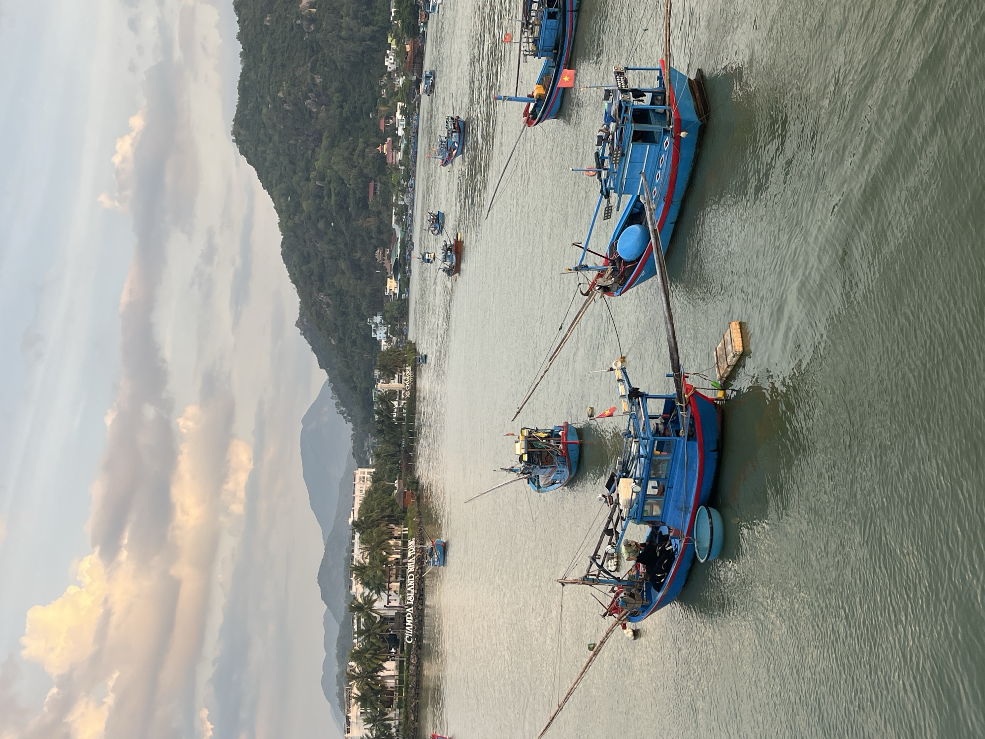 The image shows the city of Bac Nha Trang Ward, Vietnam, with a waterway occupied by blue and red fishing boats. The boats are scattered across the water, some clustered together, while others are more isolated. Mountains are visible in the background, alongside buildings and trees lining the shore. The sky is partly cloudy.