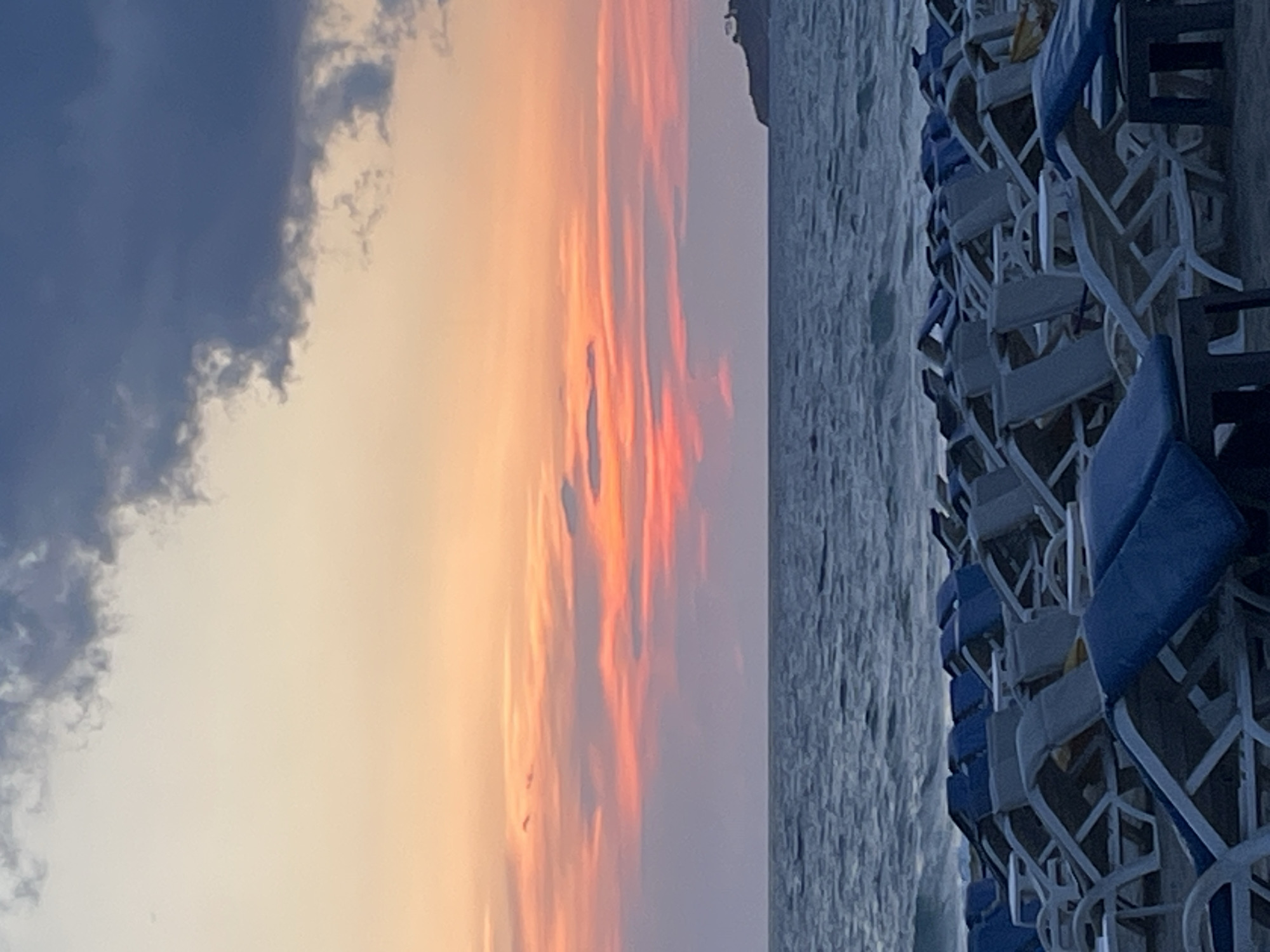 The image captures a beach scene in Alanya, Turkey, likely during sunset. The sky displays a gradient of colors, ranging from dark blue/gray clouds on the left to orange and pink hues in the center, suggesting the sun is setting. A body of water, presumably the Mediterranean Sea, is seen with gentle waves. The right side of the image shows rows of empty beach chairs, stacked closely together. The chairs are mostly white with blue cushions. The scene is generally calm, with no immediate signs of human activity visible.