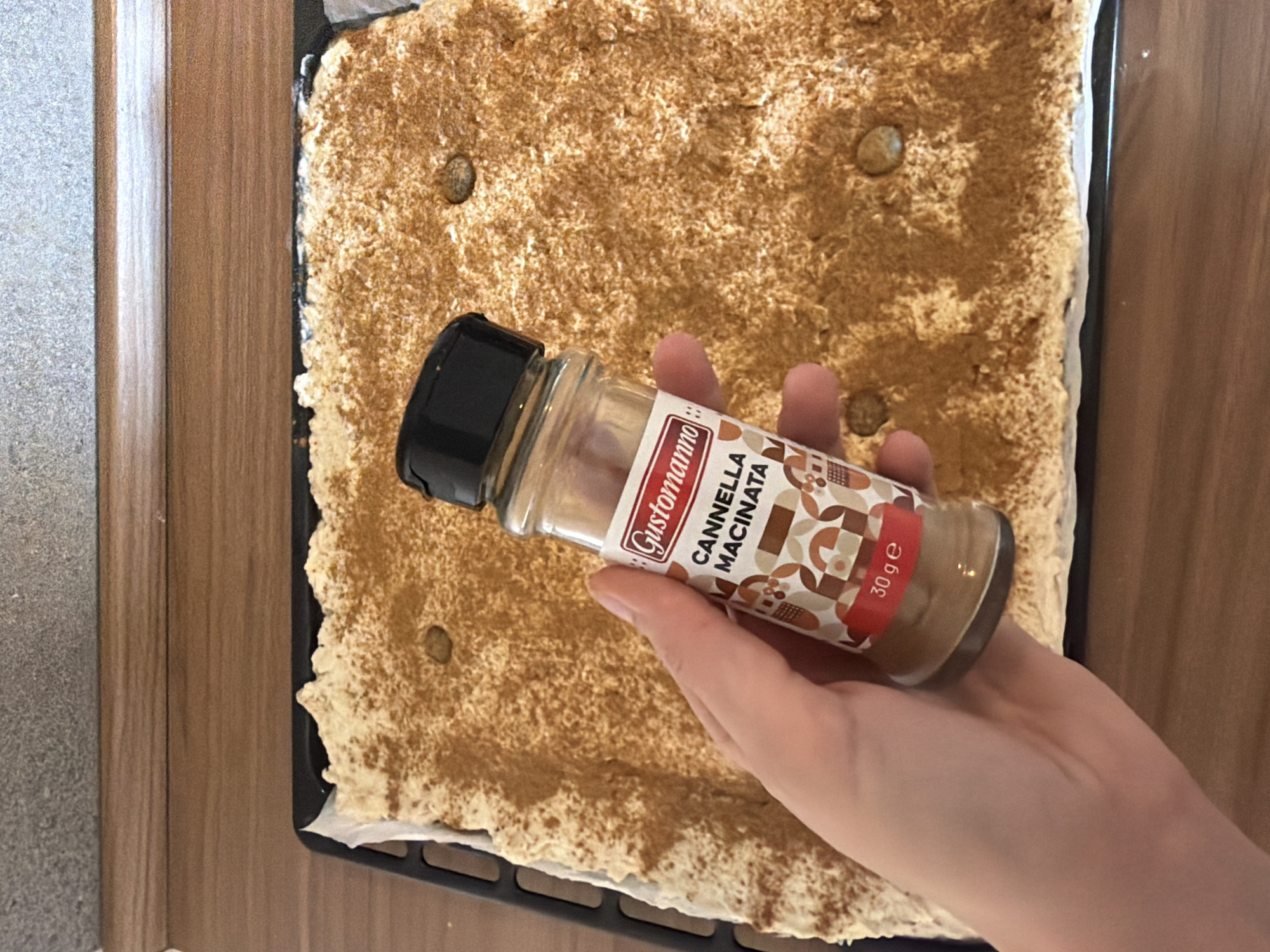 A hand holds a "Gustonanno Cannella Macinata 30g" cinnamon spice jar over a baking sheet. The baking sheet contains a light-colored dough or batter dusted with cinnamon. The scene suggests someone is preparing a dessert or baked item. The product is from Catania, Italy. The environment is a kitchen.