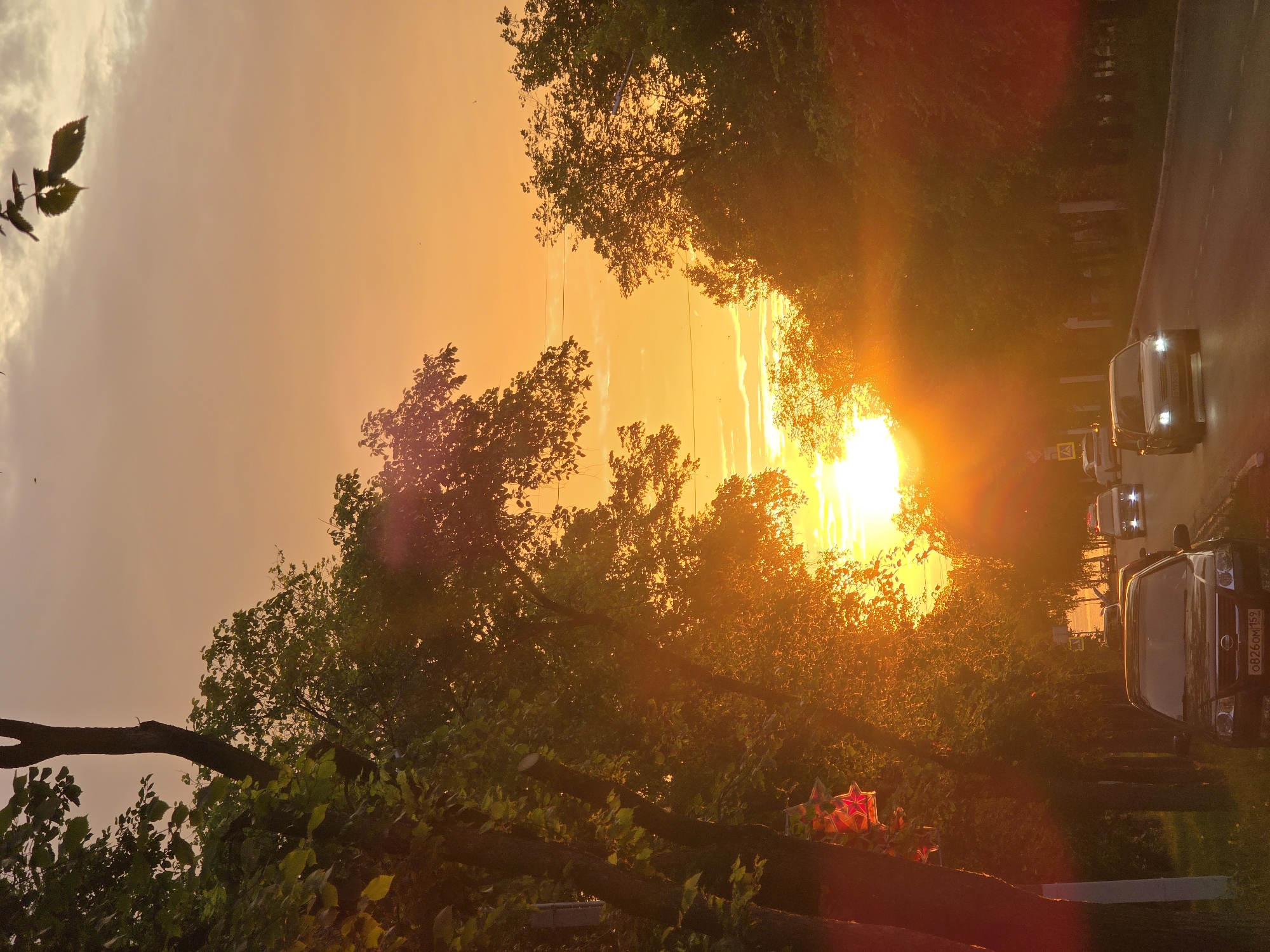 The image depicts a street scene at sunset in Chaikovsky, Russia. The sun is setting behind trees, casting a warm orange glow over the sky and illuminating the scene. Several cars are on the road, and trees line the sides of the street. There are also some decorative red stars hanging from a pole on the left side of the image. The overall impression is one of a peaceful evening in a city.