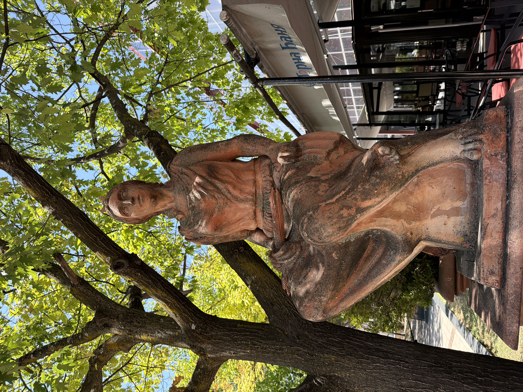 A terra cotta statue of a seated woman, partially covered in moss, stands in front of a large tree with green leaves. The statue is reddish-brown and detailed, with a crown on its head and draped clothing. Its posture is relaxed, with legs crossed. Behind the statue and tree, a building with glass windows and outdoor seating is visible. A white awning with text in Cyrillic script, "Горна Баня," is stretched over the seating area. The scene appears to be an outdoor seating area of a restaurant or cafe in Lovech, Bulgaria, with the statue serving as a decorative element. The lighting suggests a sunny day.