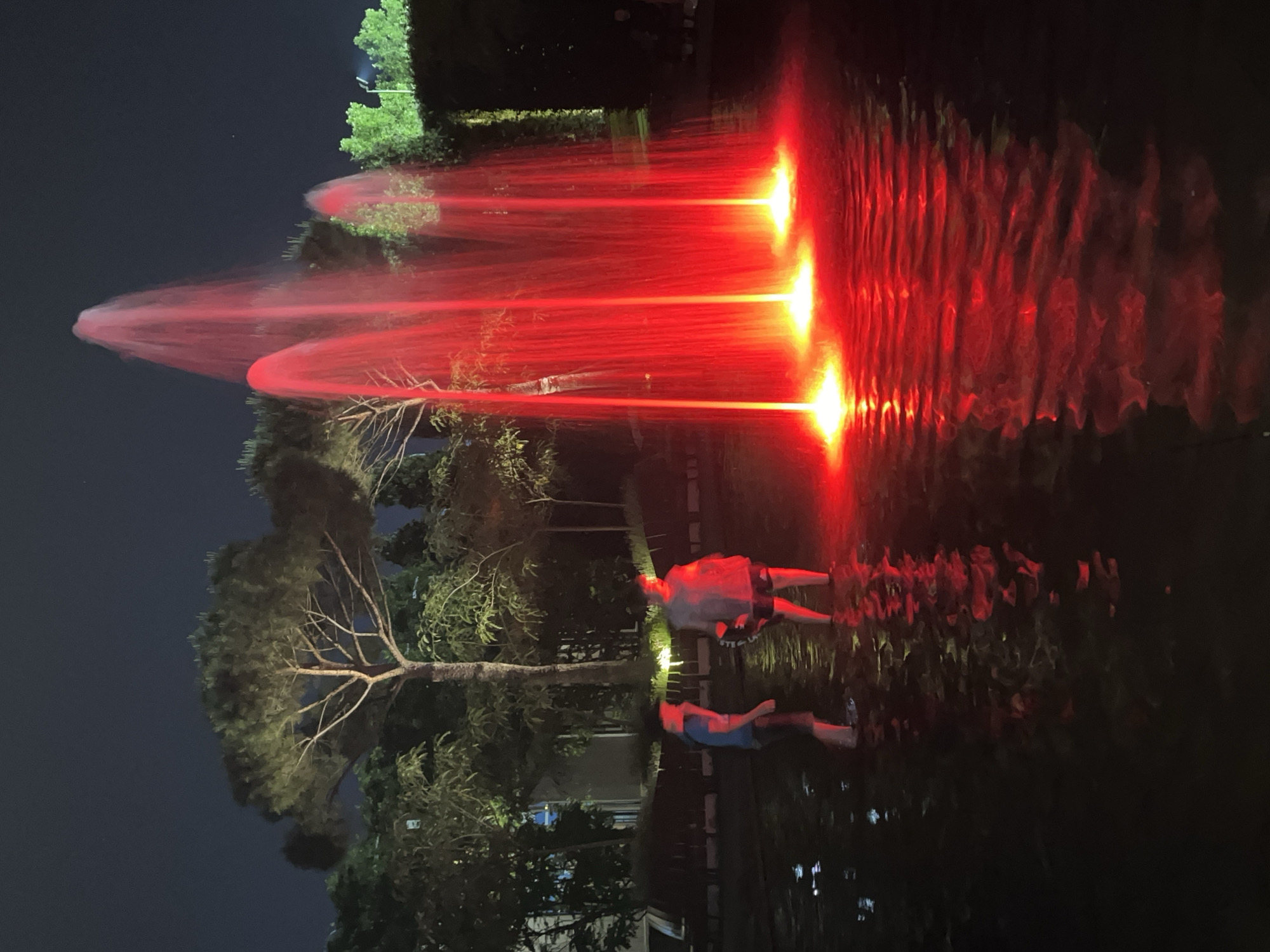 The image depicts a nighttime scene with a fountain illuminated by red lights, casting a vibrant glow on the surrounding water and foliage. Two individuals, appearing to be children or young teenagers, are standing in the shallow water of the fountain, facing away from the camera. The water is dark, reflecting the red light from the fountain, creating a mesmerizing visual effect. In the background, there are several trees, including a prominent pine tree, and the silhouettes of buildings and possibly streetlights are visible. The overall atmosphere is serene and somewhat mystical, with the vibrant red of the fountain contrasting against the dark, nocturnal environment. This scene is located in the city of Krasnodar, Russia.